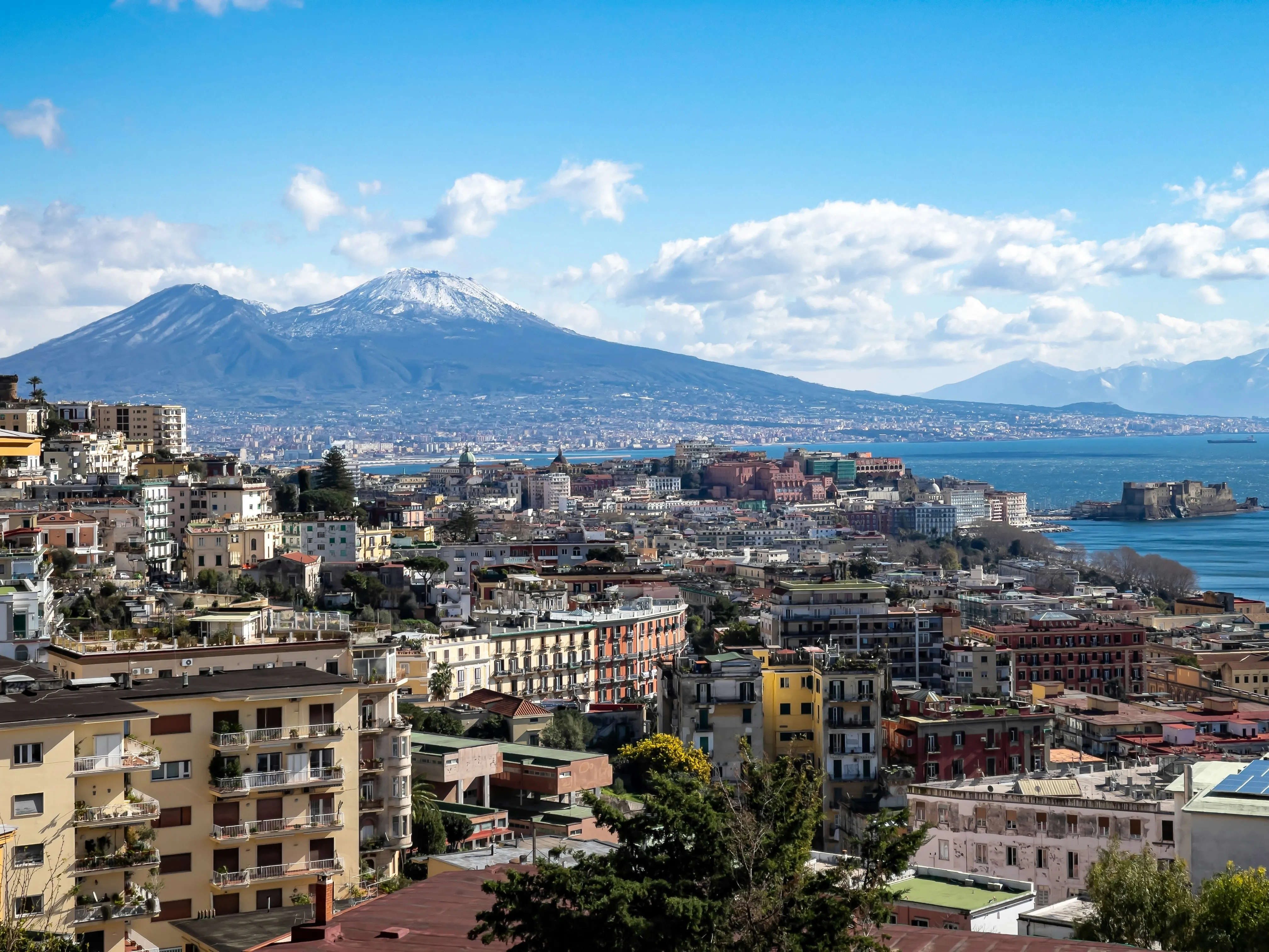 Scenic view of Naples city with colorful buildings, blue sea, and snow-capped Mount Vesuvius in the background.
