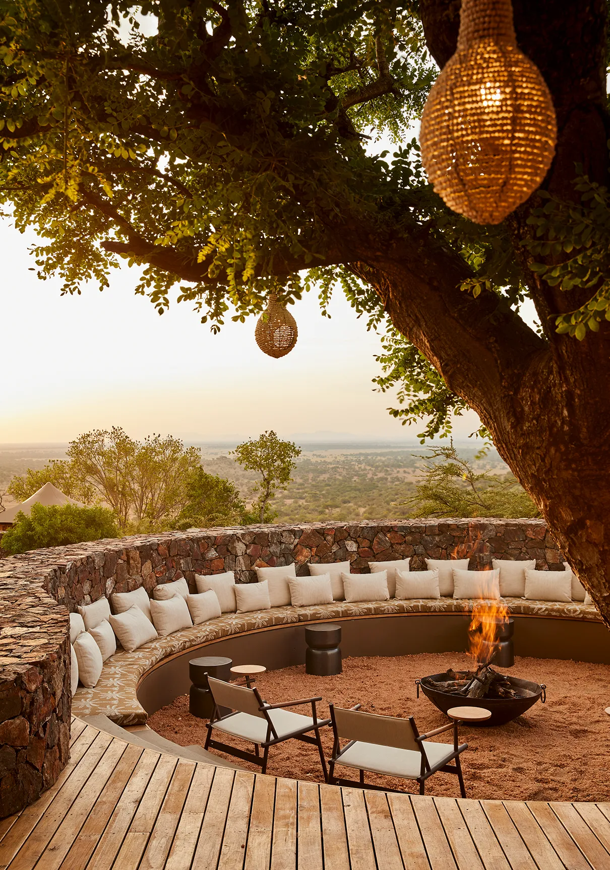 Sunken outdoor fire pit with curved seating beneath a large tree, overlooking a scenic savannah at sunset.
