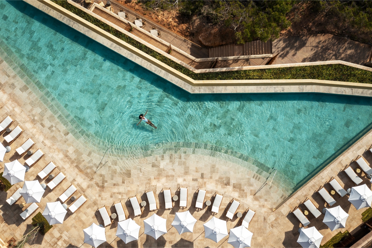 Aerial view of a person swimming in a long, narrow pool surrounded by lounge chairs and umbrellas.