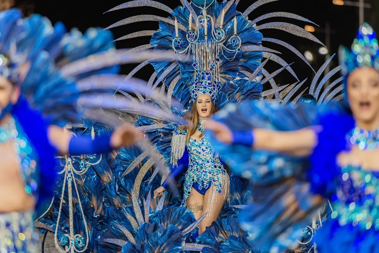 Performer in an elaborate blue feathered carnival costume surrounded by dancers in matching blue outfits.