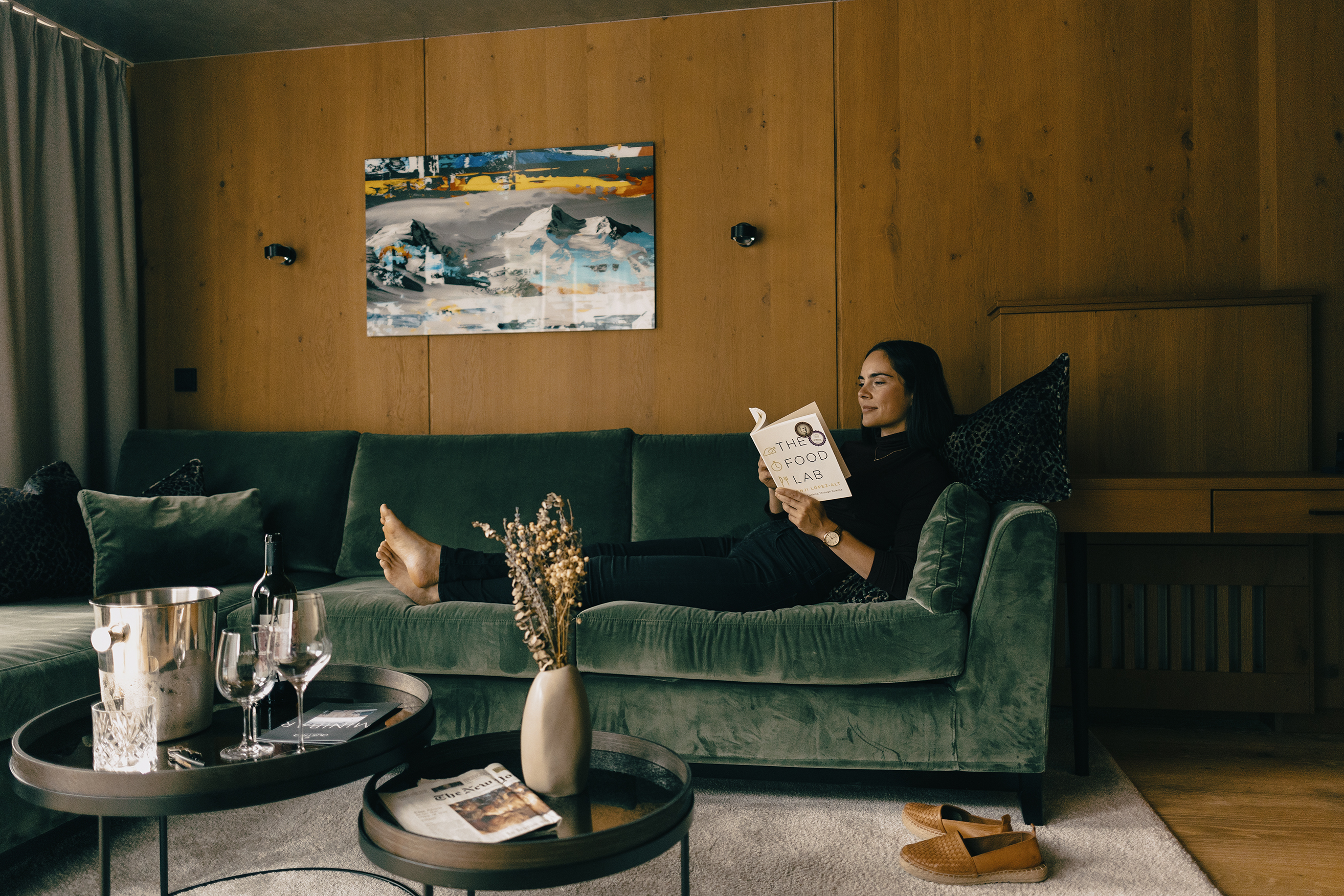 A person relaxing on a green velvet sofa reading in a cosy, modern living room at the Gourmet and Wine Hotel Austria, with wooden walls, abstract art, and a wine-set coffee table