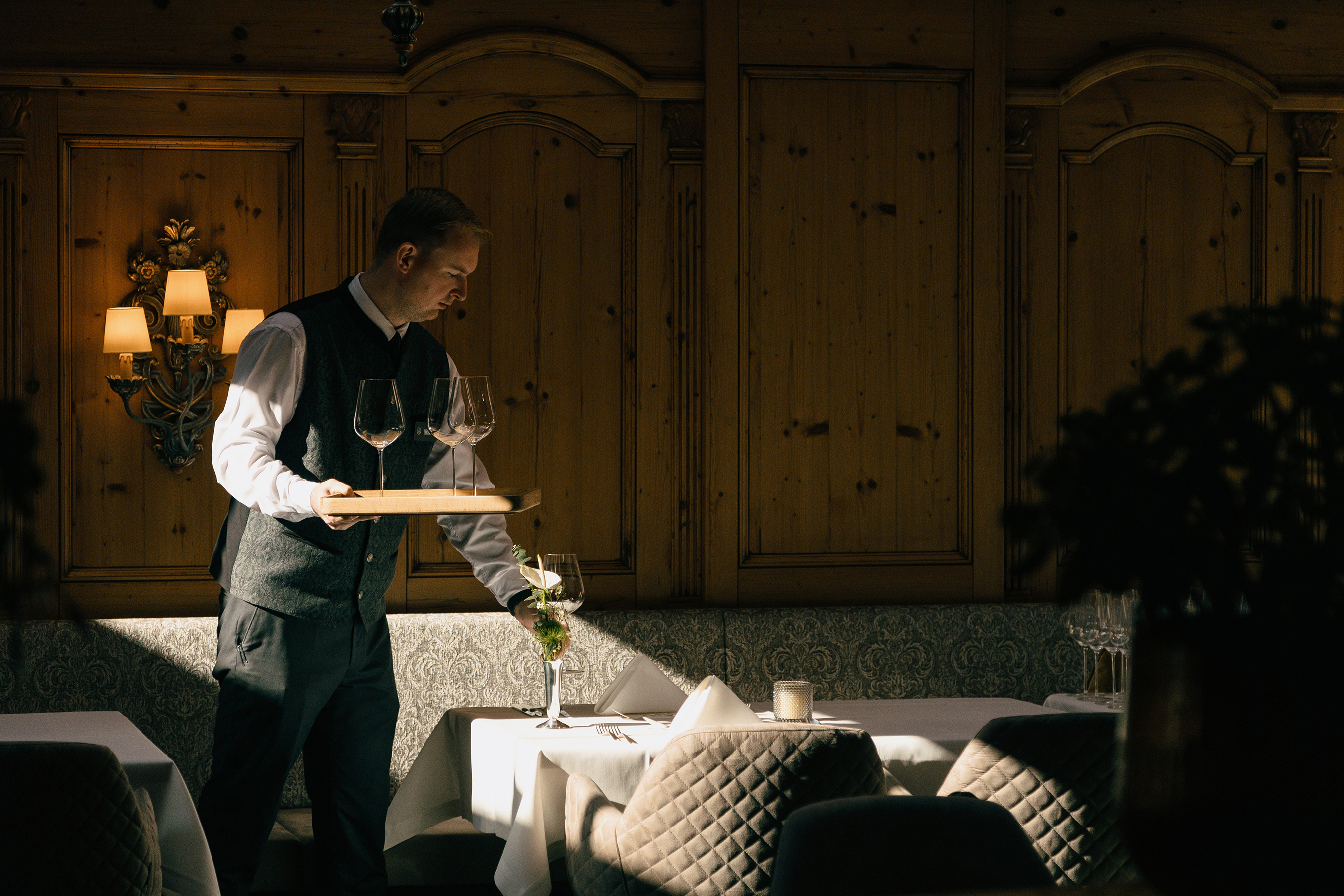 A waiter in formal attire carrying a tray of wine glasses in a warmly lit, elegant dining room at the Gourmet and Wine Hotel Austria, featuring wooden paneling and neatly set tables