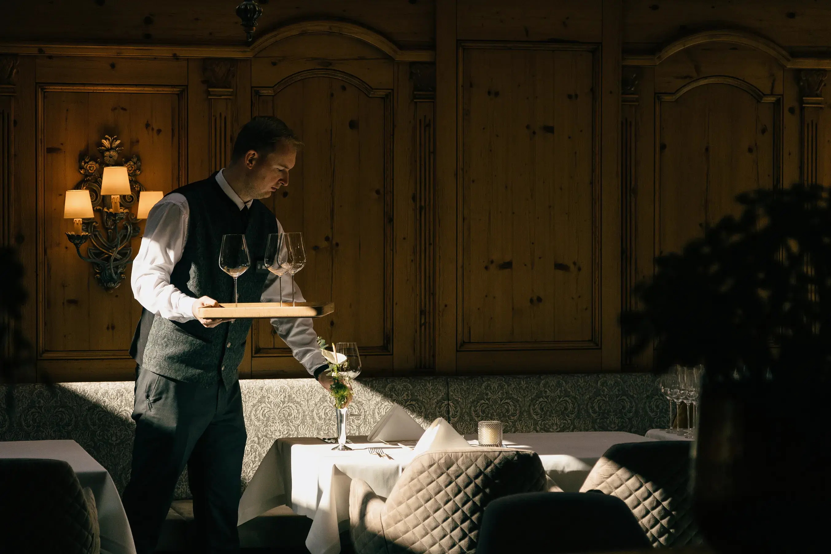 A waiter in formal attire carrying a tray of wine glasses in a warmly lit, elegant dining room at the Gourmet and Wine Hotel Austria, featuring wooden paneling and neatly set tables