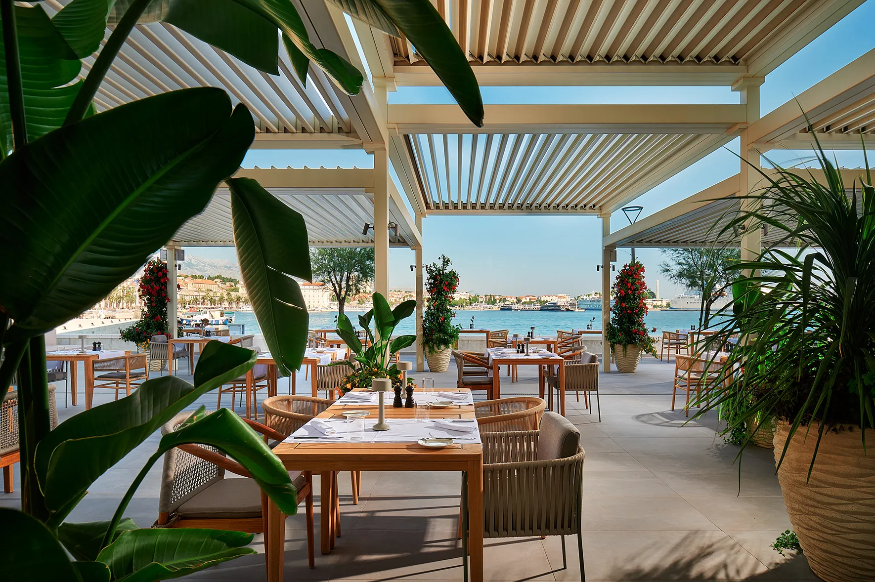 Open-air waterfront restaurant with modern seating, lush plants, and harbor views.