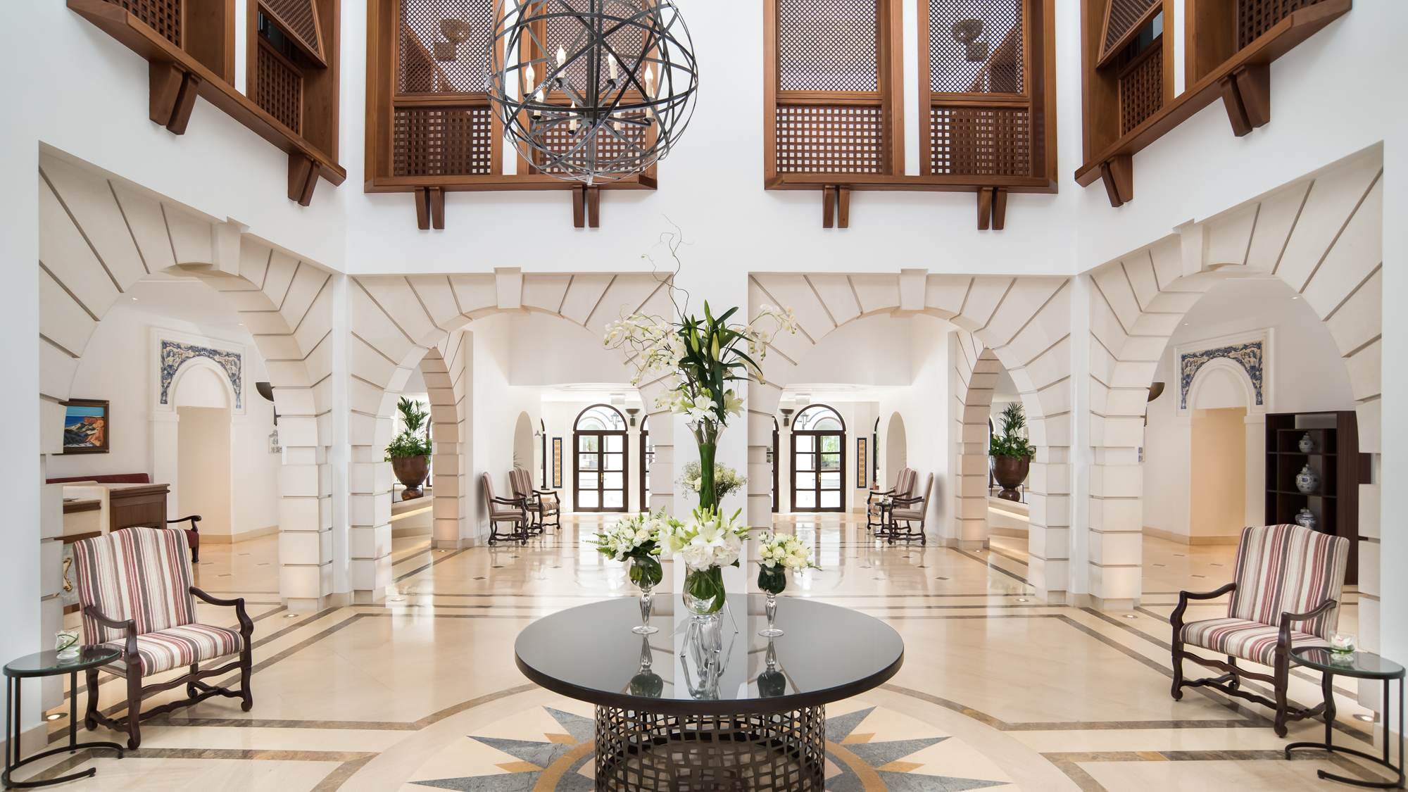 A spacious, elegant interior with high ceilings, wooden balconies, arched doorways, and a central table with floral arrangements, surrounded by striped chairs and intricate tile floorin