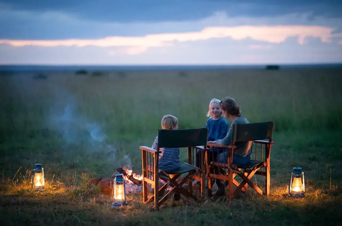 A family of three sitting around a campfire at sunset in the African bush with lanterns positioned around 