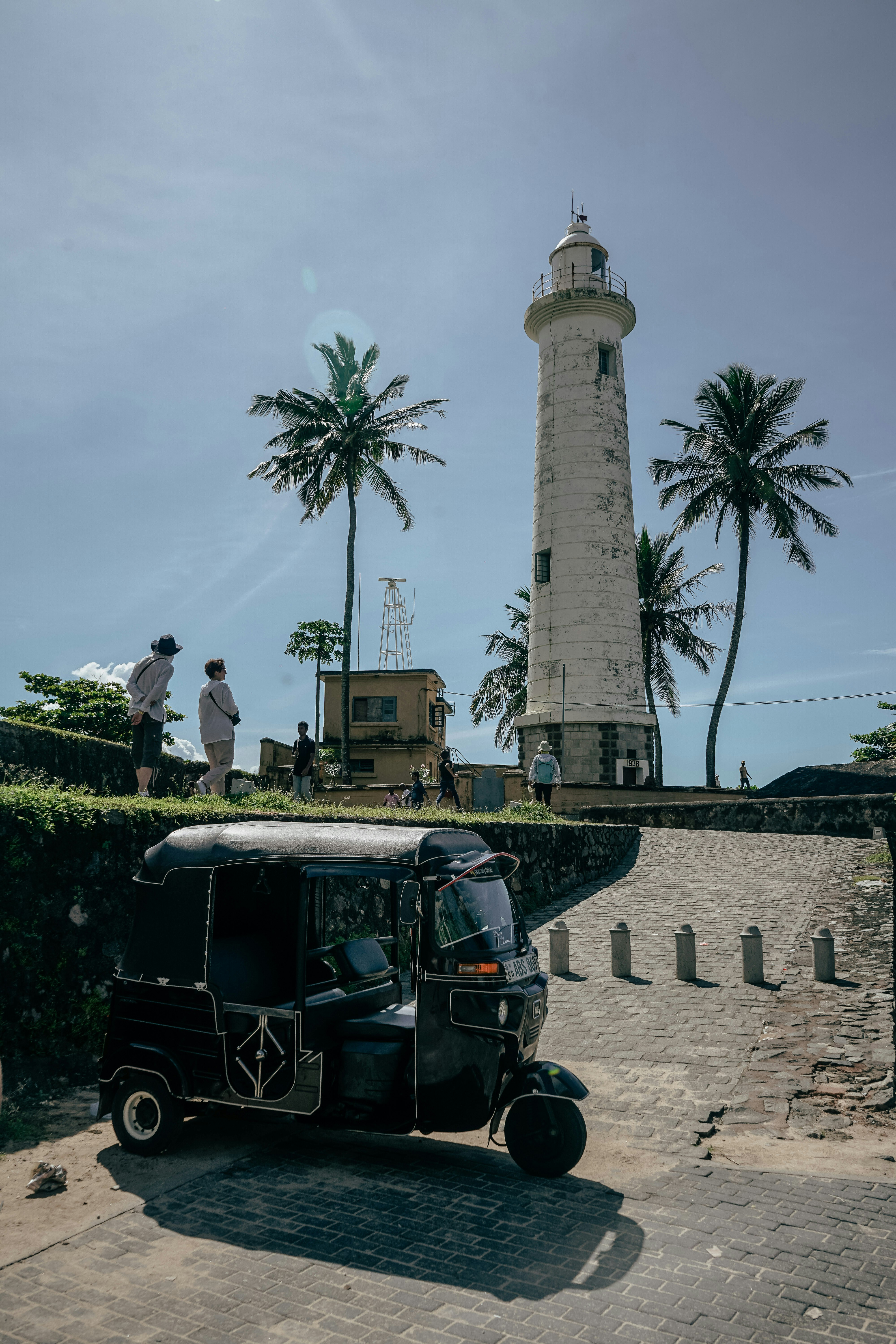Historic coastal lighthouse in Galle surrounded by palm trees with a tuk‑tuk parked on a cobblestone path and visitors exploring the area under a bright sky.