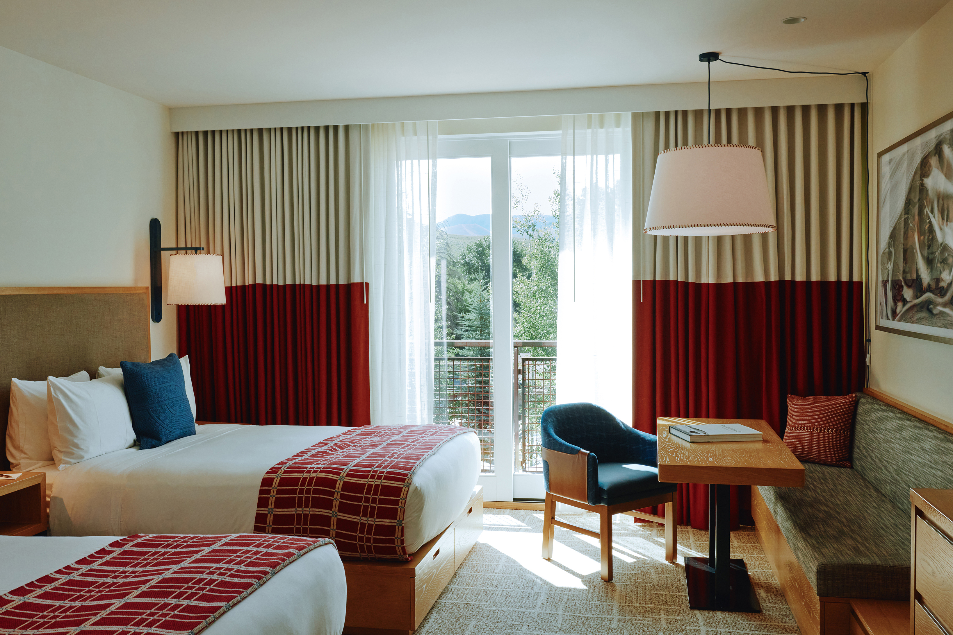 A hotel room at Limelight Ketchum with two double beds dressed in red and white patterned blankets, a window with curtains, a small table and chair, and a bench seat