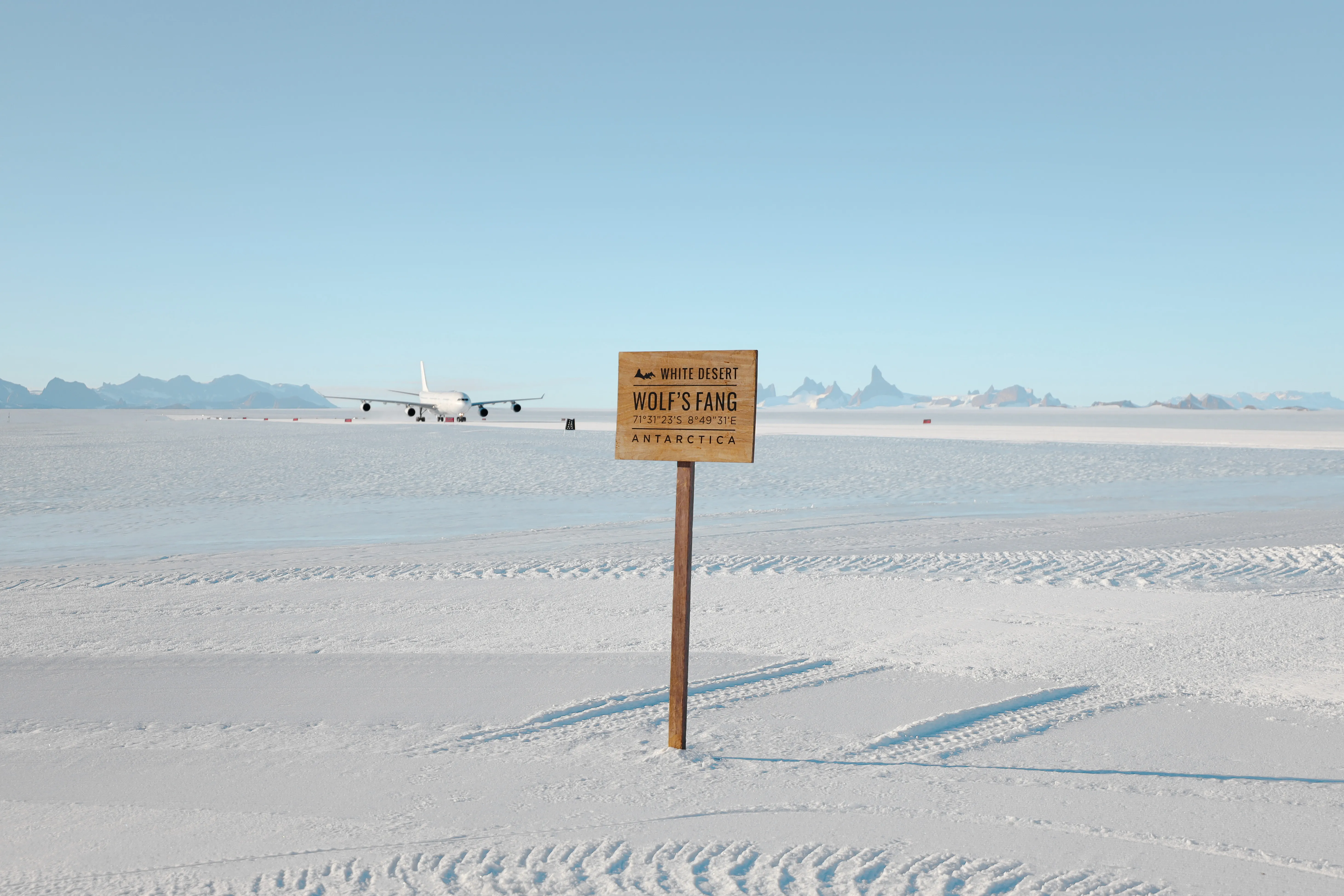 Sign marking Wolf’s Fang runway in Antarctica with an aircraft parked on the snowy ice landscape under a clear blue sky.