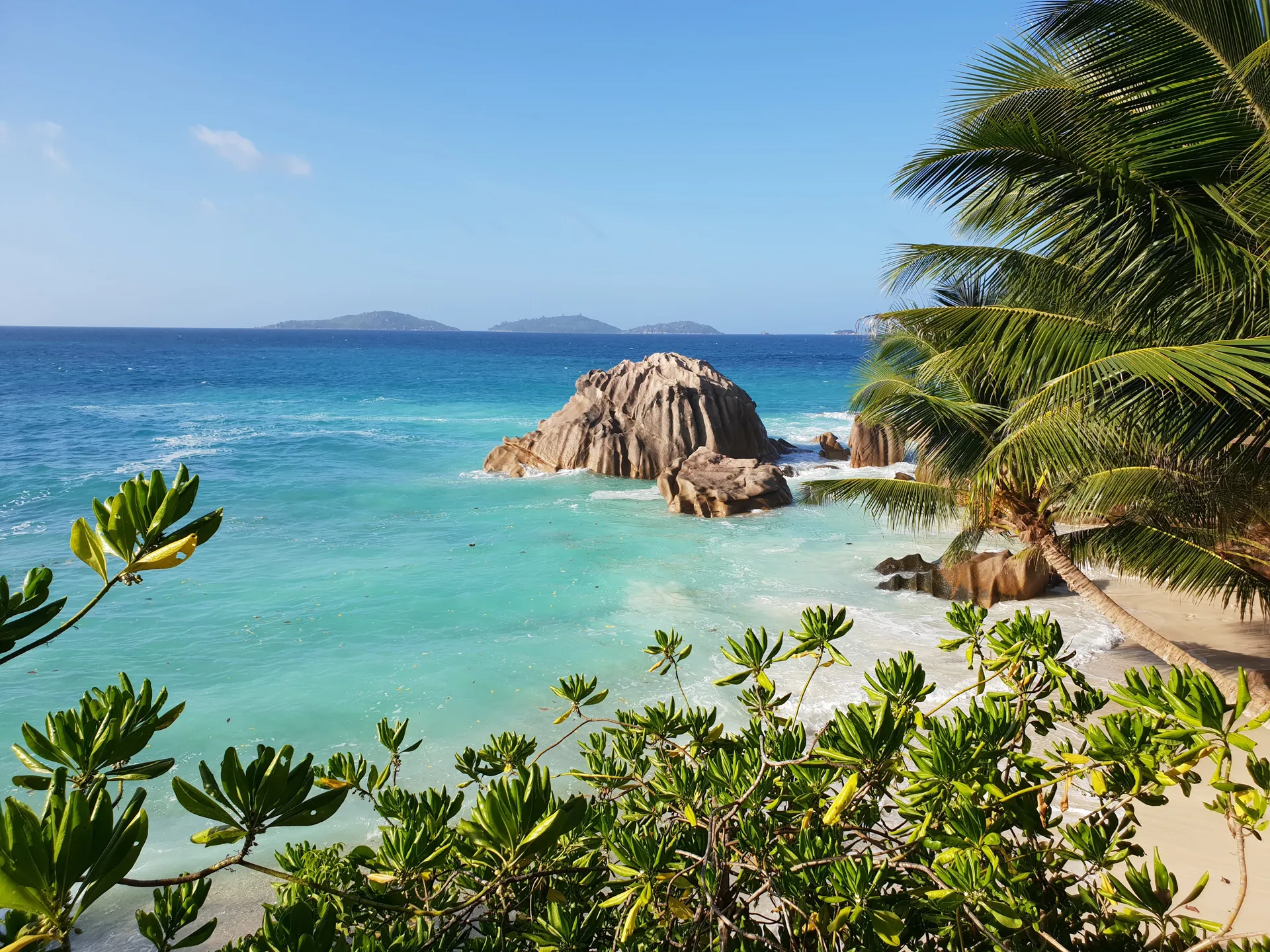Tropical beach with turquoise waves, granite rock formations and lush greenery under a clear blue sky in the Seychelles.