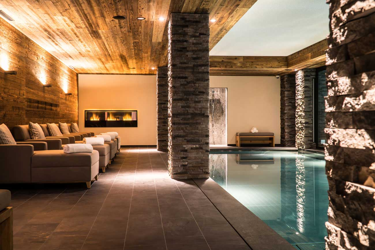 Tranquil indoor spa with a heated pool, stone pillars, loungers, and a softly lit fireplace.