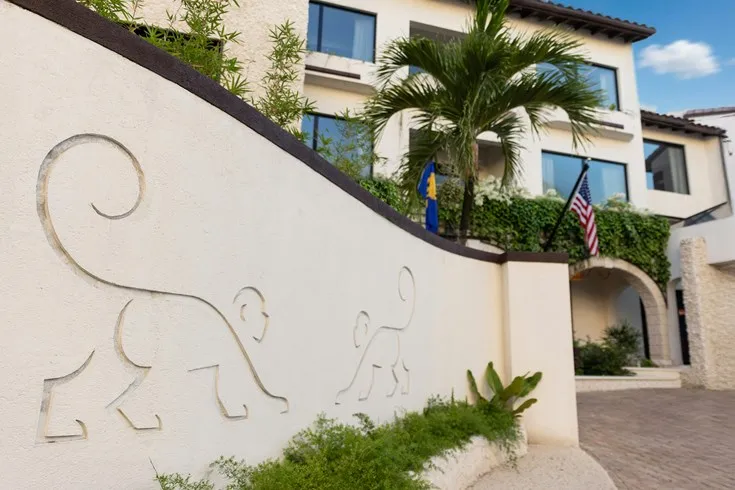 Exterior of a resort-style building with monkey silhouettes carved into a curved wall, tropical landscaping, and flags near the entrance.