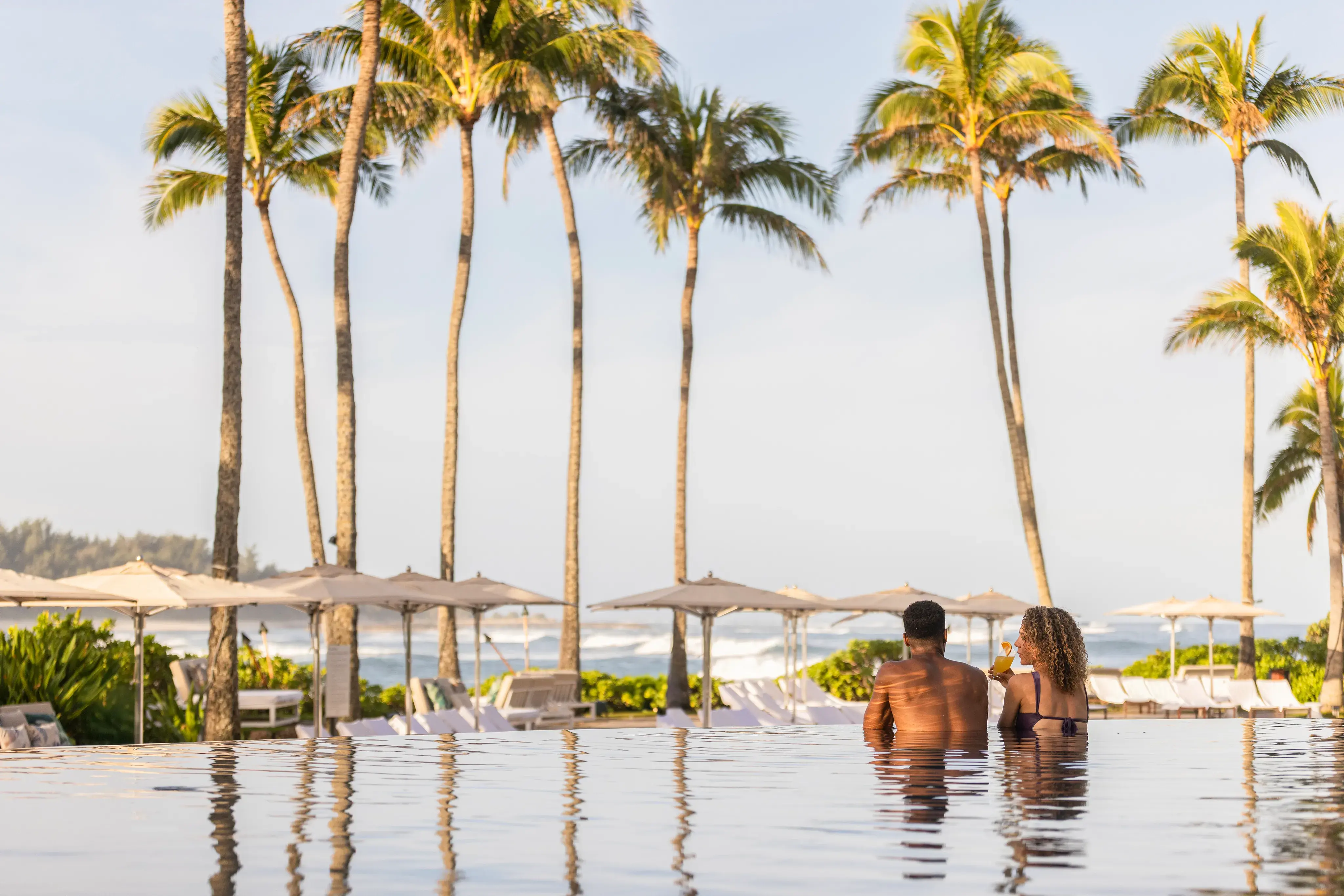 Two guests looking over the edge of the adults only infinity pool at Ritz-Carlton Turtle Bay, fronted by palm trees and sun umbrellas