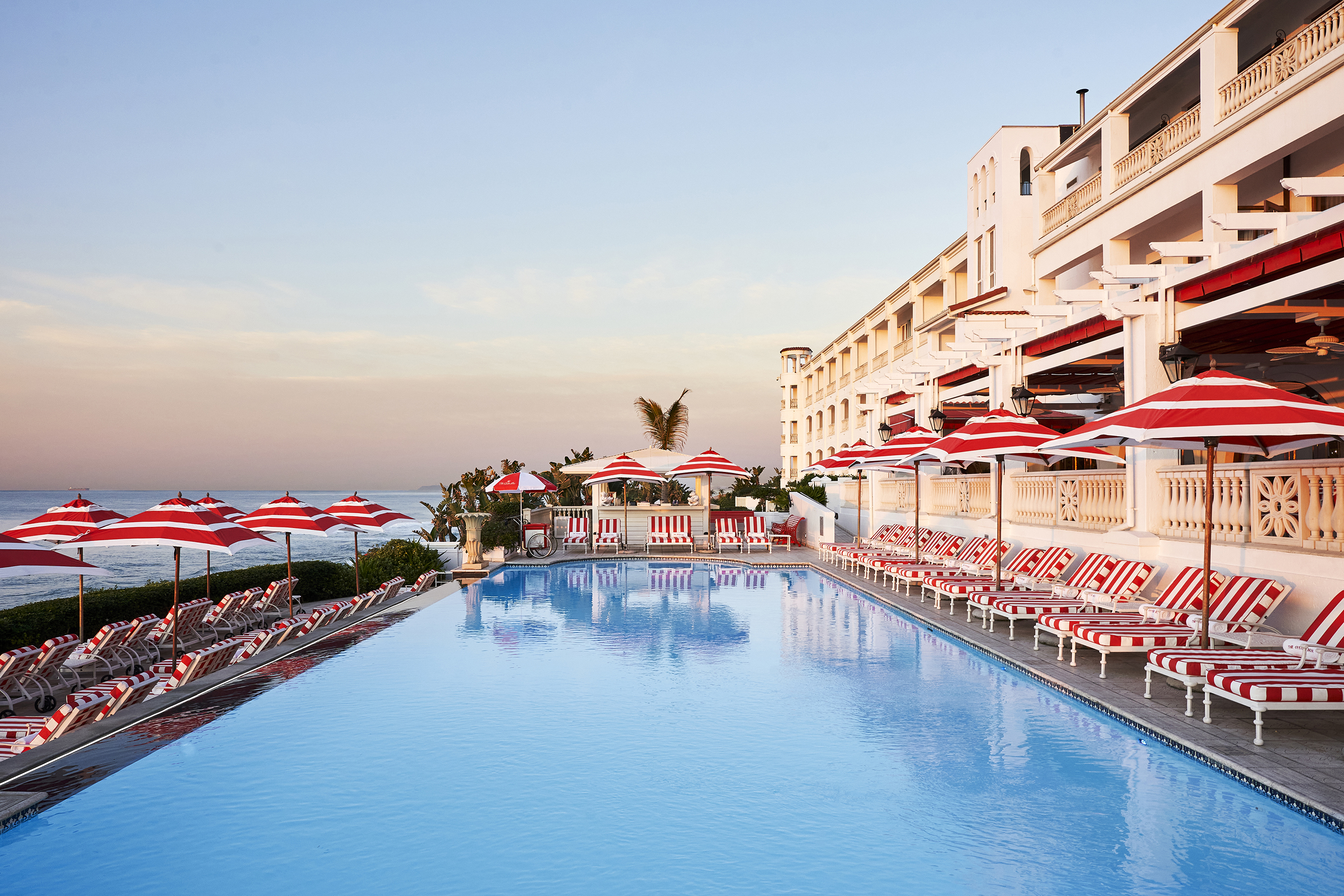 The outdoor pool at The Oyster Box lined with red and white sun loungers and the side of the hotel facing the ocean