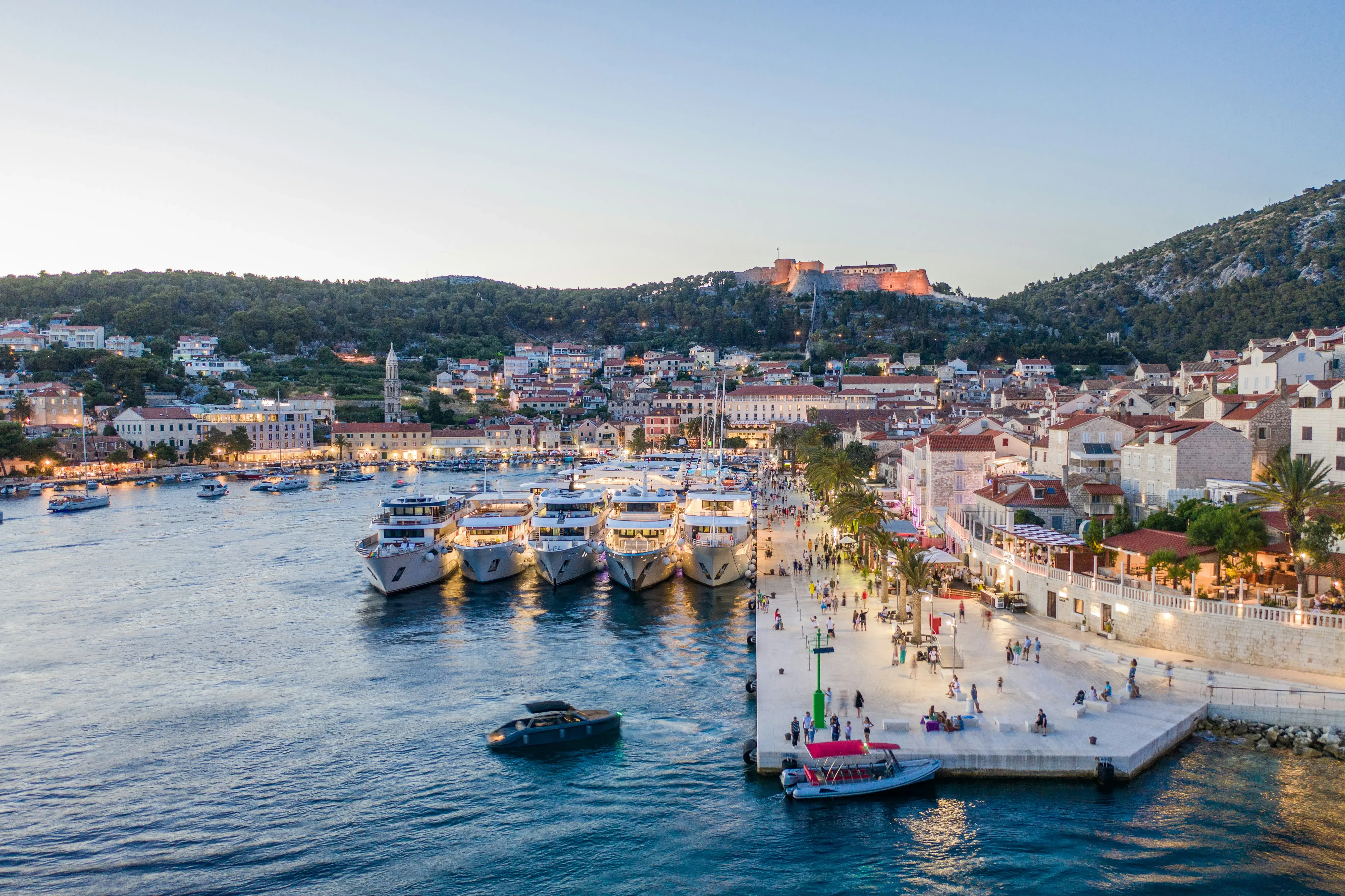 Hvar's port lit up at dusk featuring yachts and a busy promenade in front of the town and fortress on a hill