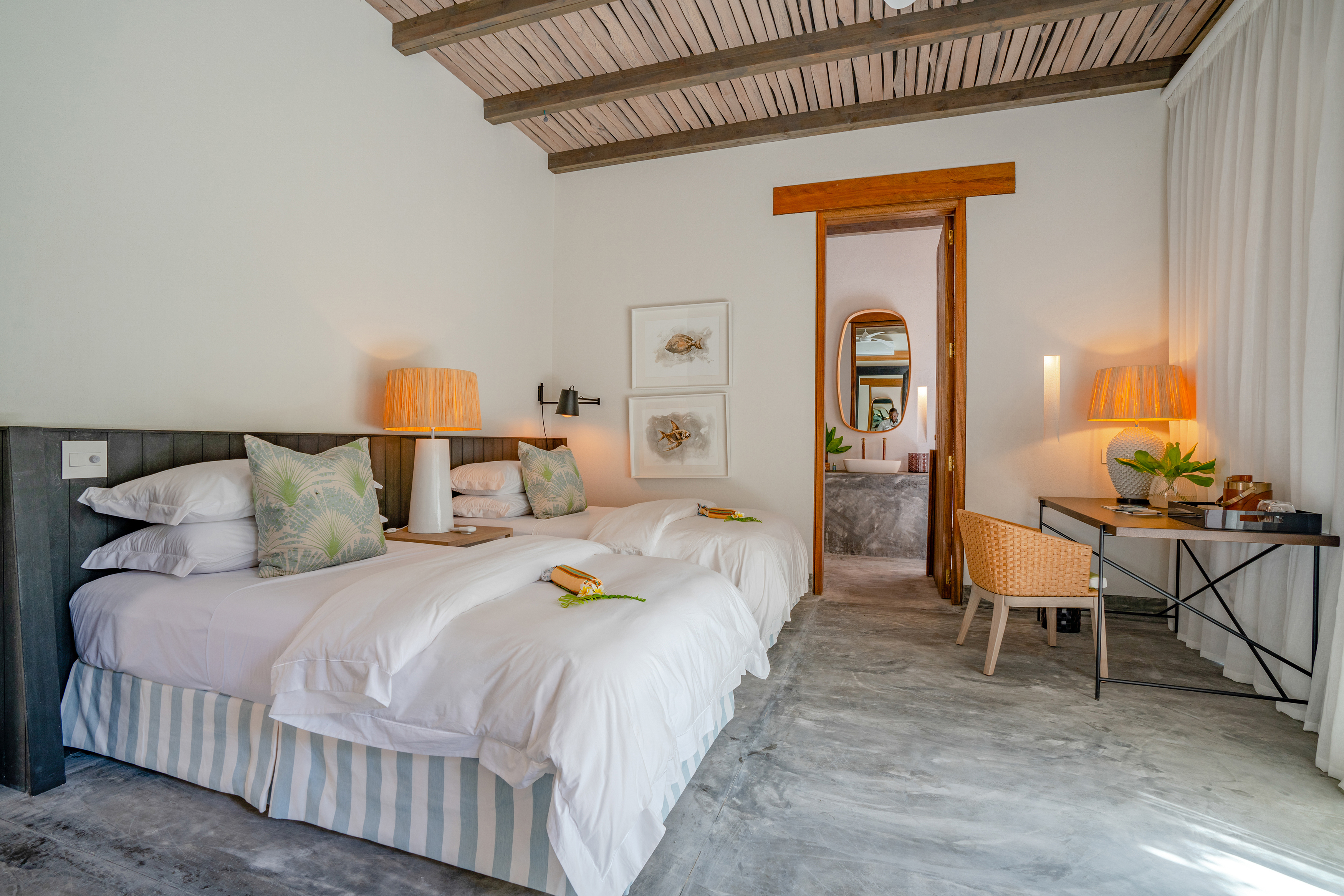 A large airy room with thatched roof featuring two plush beds and furnishings, with a glimpse of a bathroom through the door