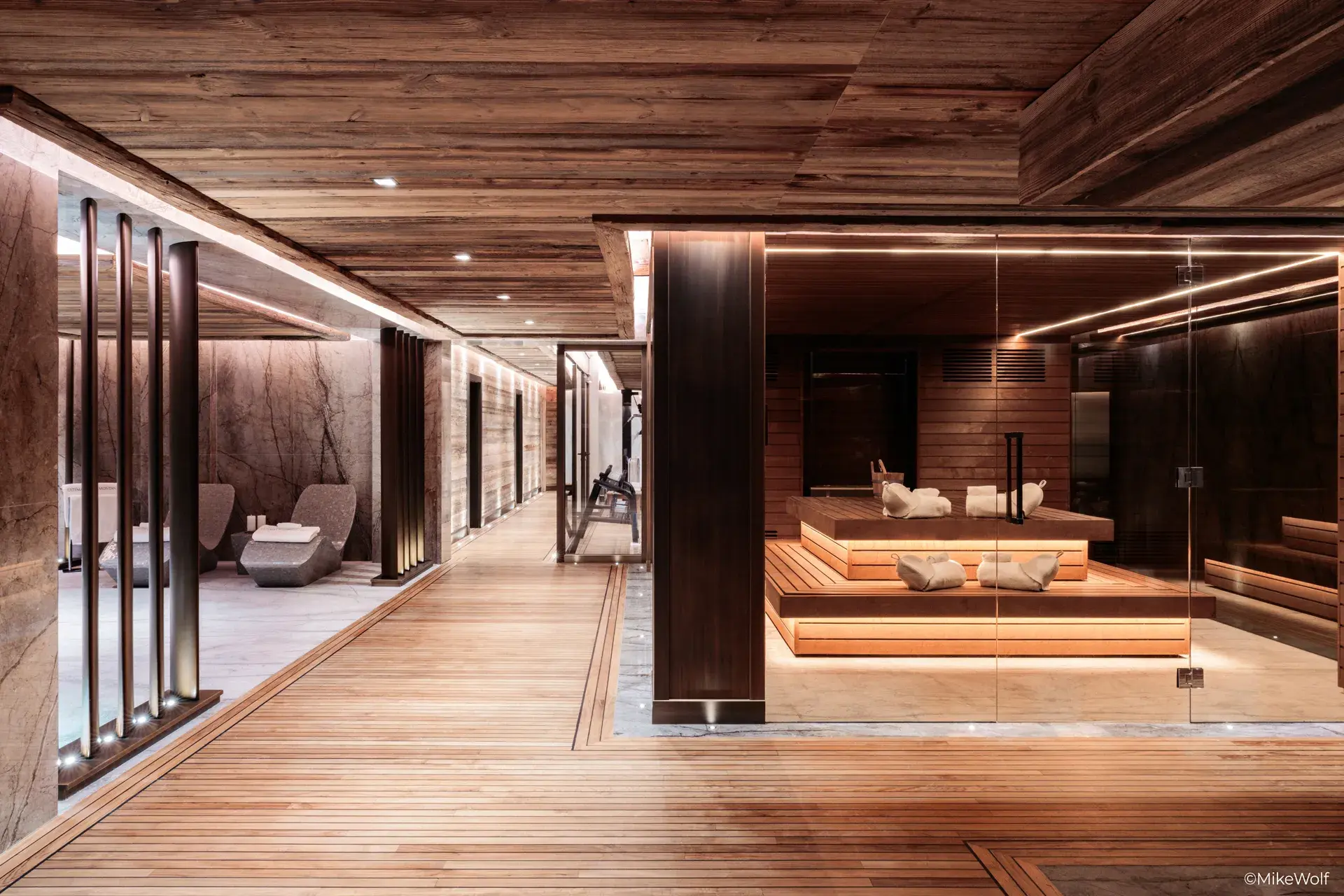 Large spa in Ultima Crans-Montana chalet featuring a large glass sauna with wooden seating opposite a relaxing marble spa lounge area with a glimpse of a gym down the corridor