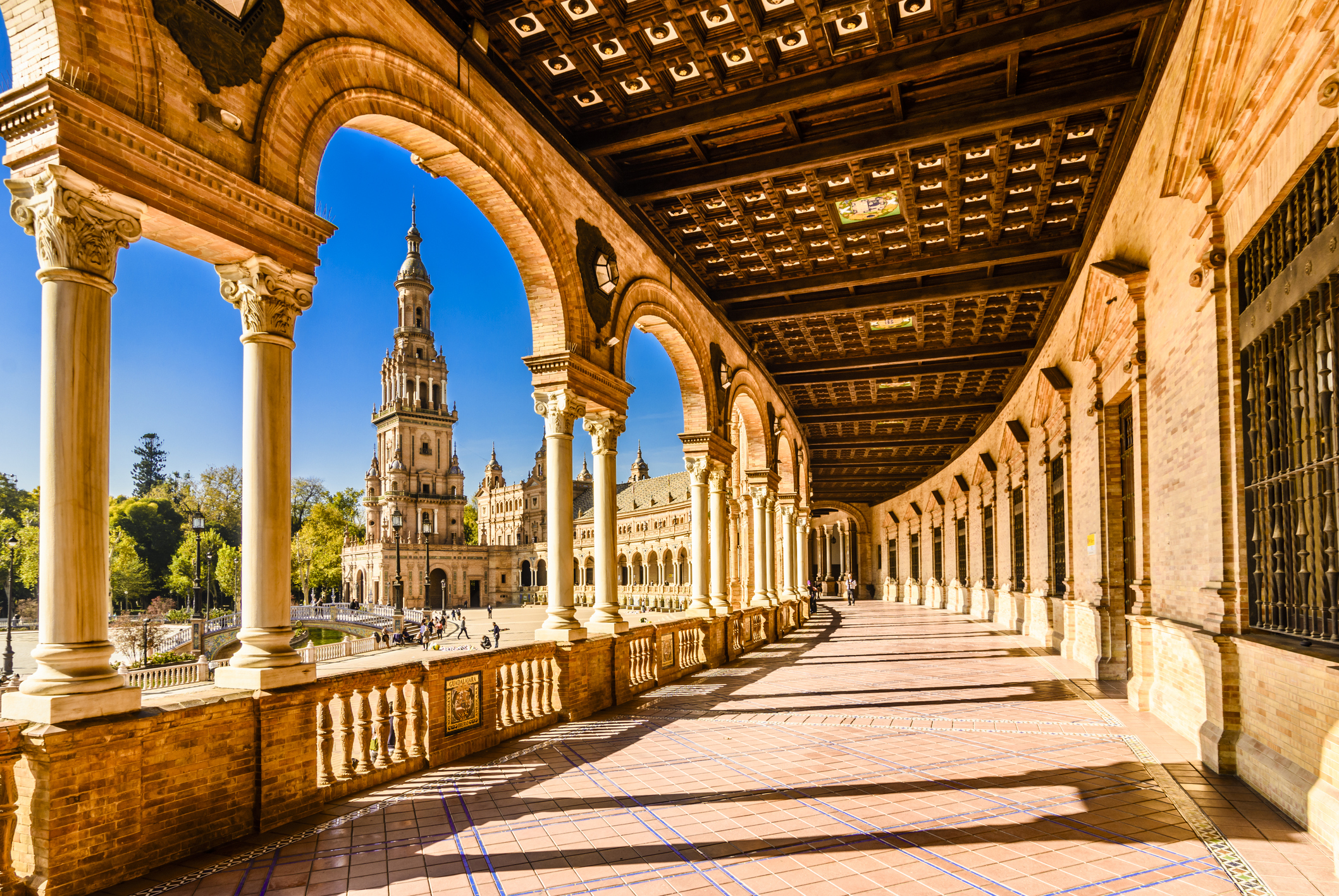 A sunlit view of the ornate arches and columns of the Plaza de España in Seville, with a clear blue sky above.