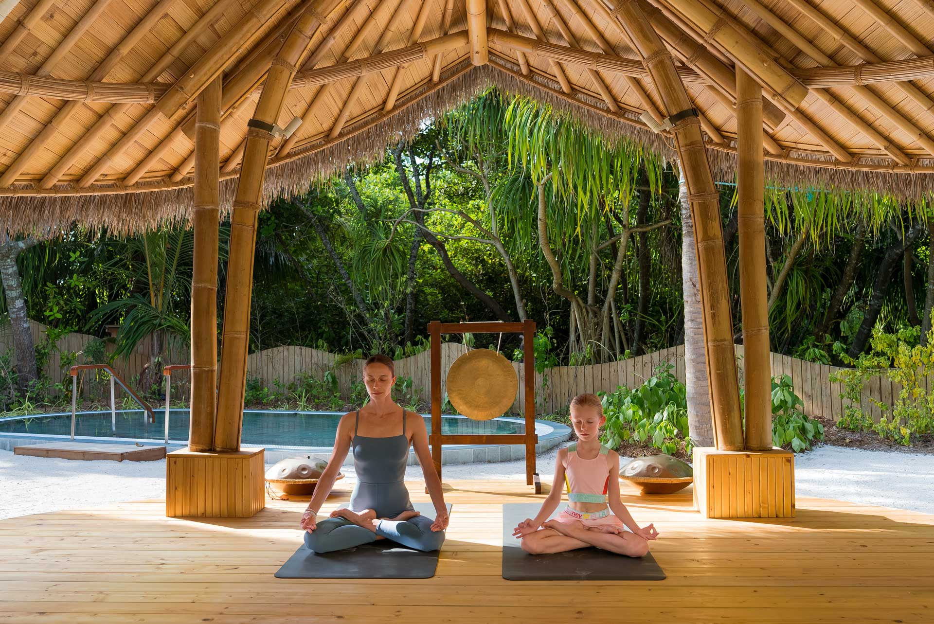 Outdoor yoga session at JOALI BEING with two guests practicing meditation beneath a bamboo pavilion surrounded by tropical greenery.