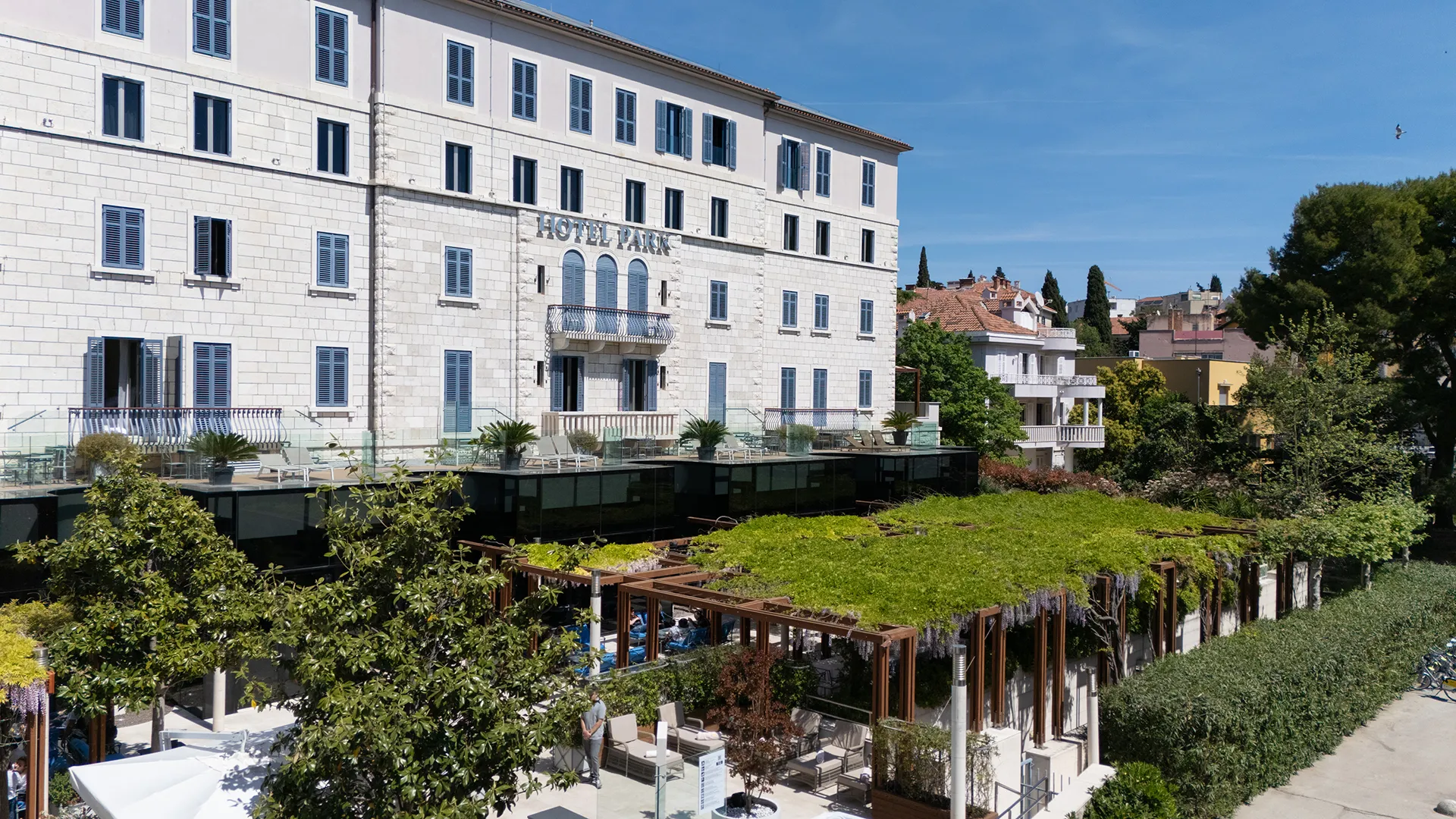 White stone hotel building with blue shutters, overlooking a landscaped terrace and garden.