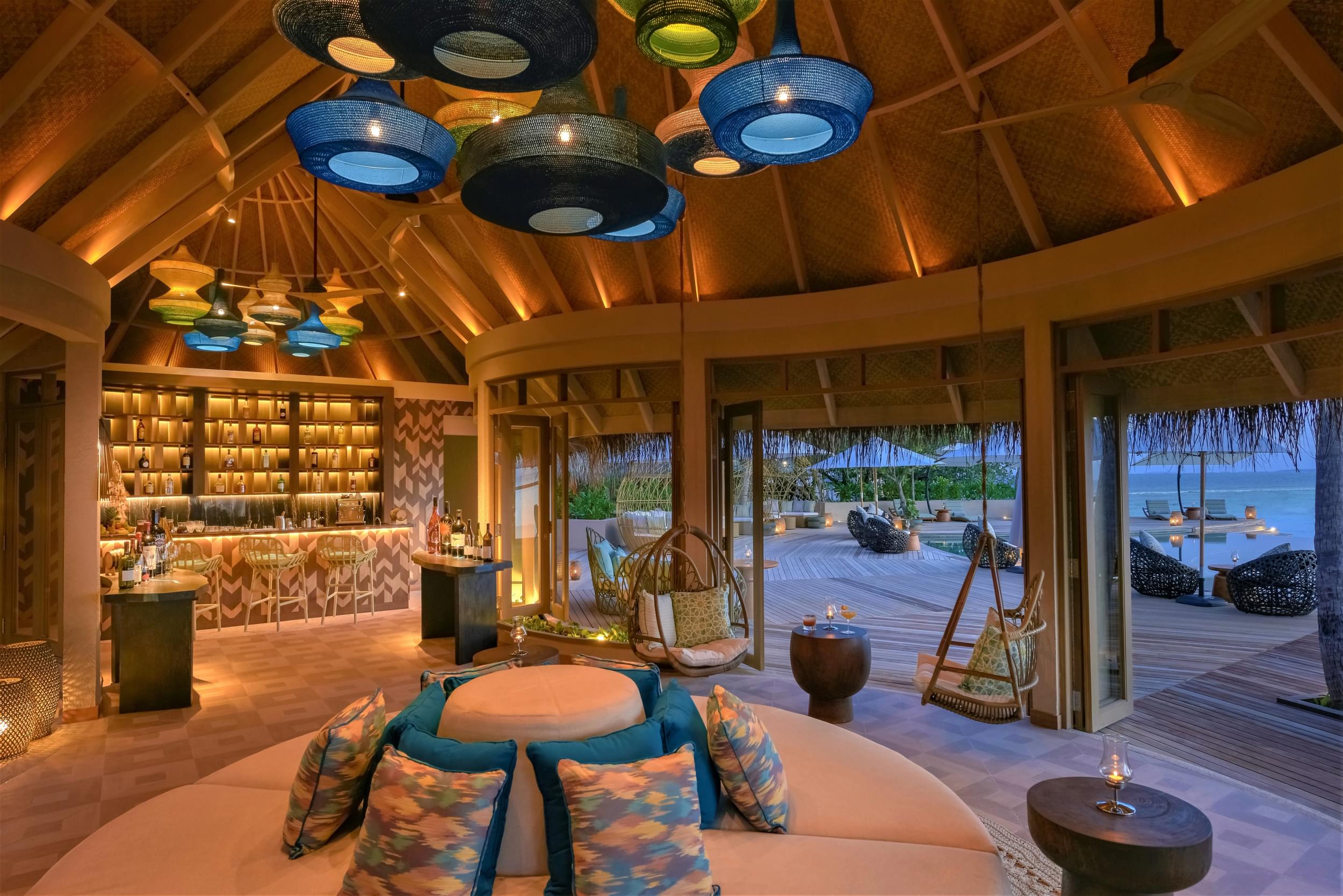 Luxurious tropical resort interior with thatched roof, elegant hanging lamps, and a view of the ocean at dusk.
