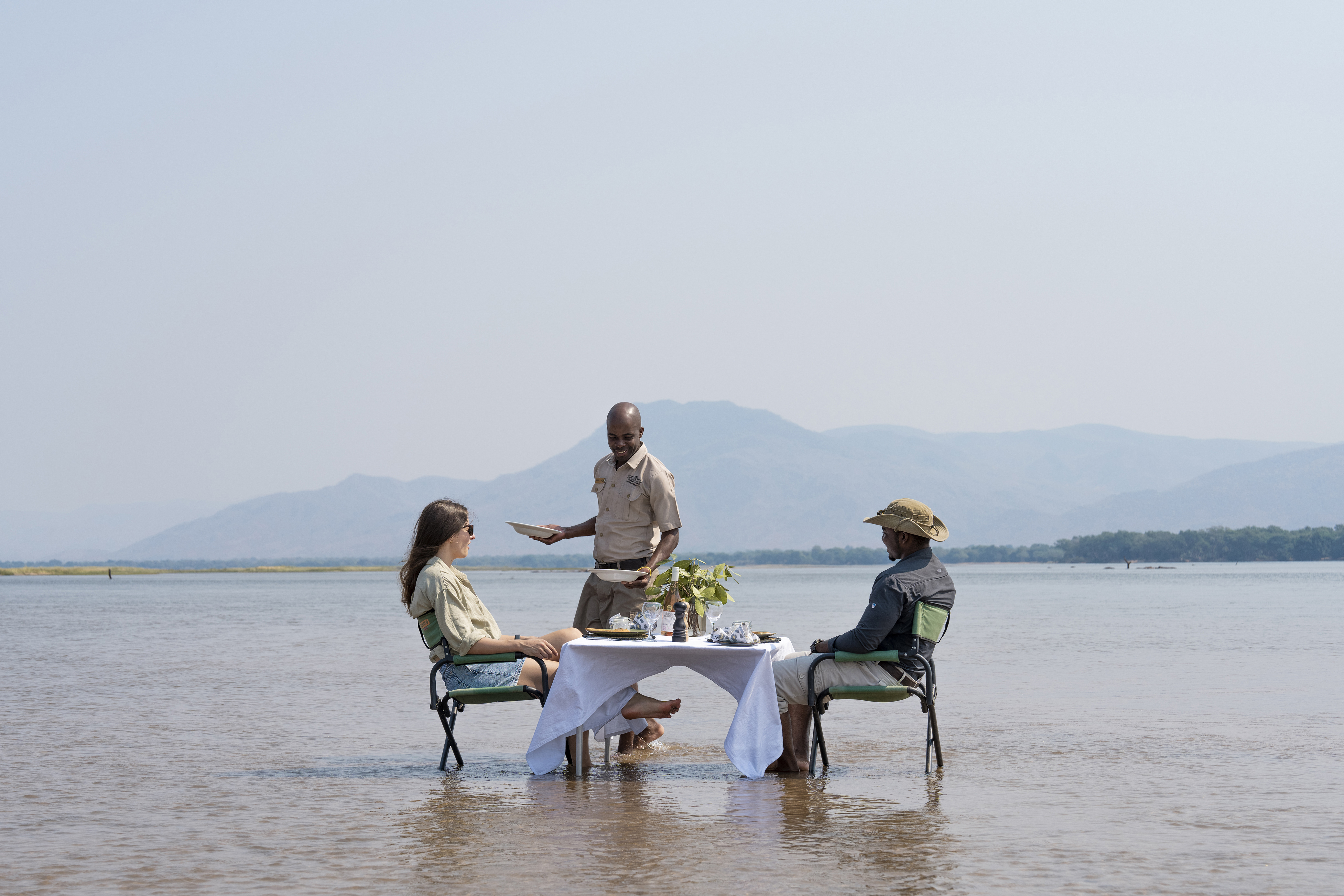 Unique dining experience with two people seated at a table in the shallow water of the Zambezi, served by a waiter, with mountains in the background.