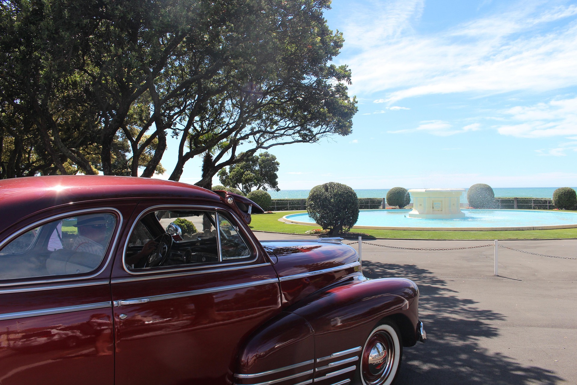 A vintage maroon car parked near a circular fountain with trimmed bushes, trees, and the ocean in the background under a bright blue sky