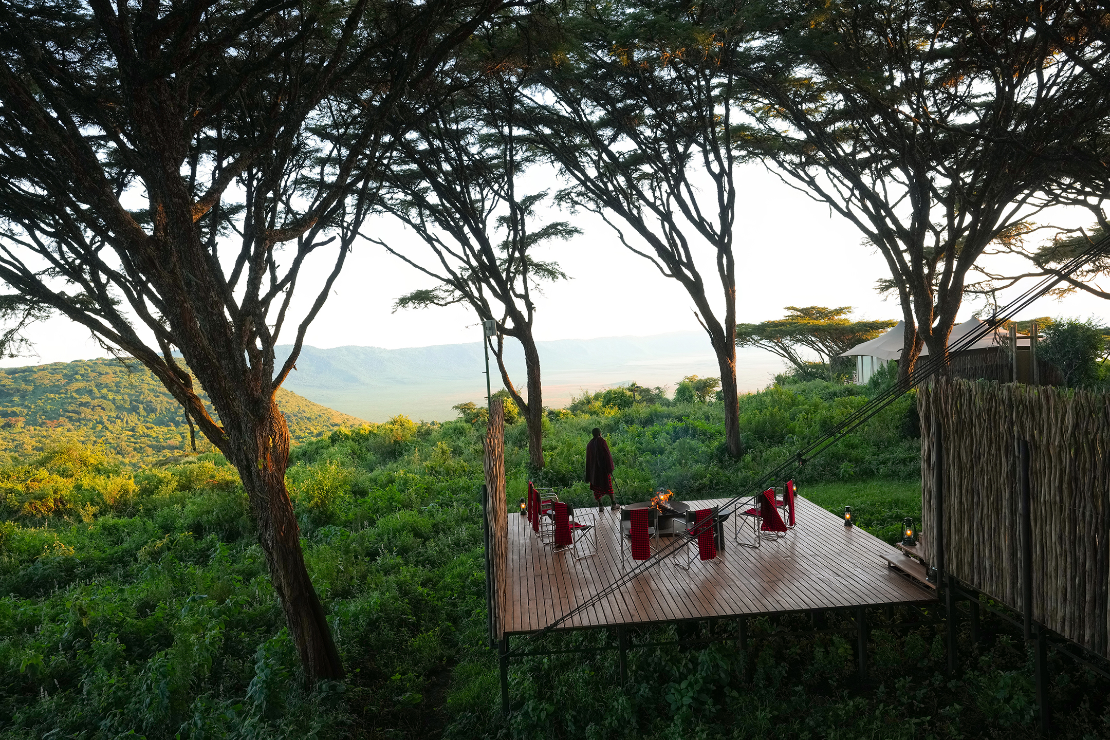 An outdoor dining set up on a platform hanging over greenery with a sweeping view of trees and the edge of the Ngorongoro Crater