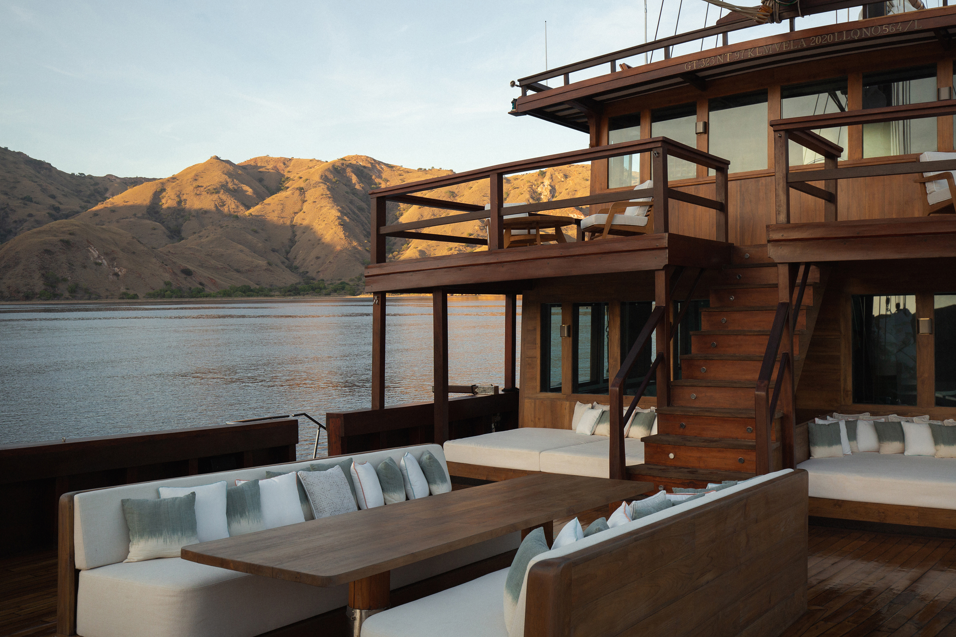 The main wooden deck of Vela yacht featuring comfy white sofas and an island in the background