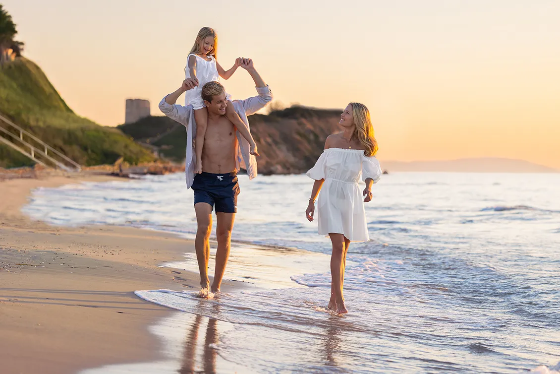 Family walking along a sandy beach at sunset with gentle waves and coastal cliffs in the background.