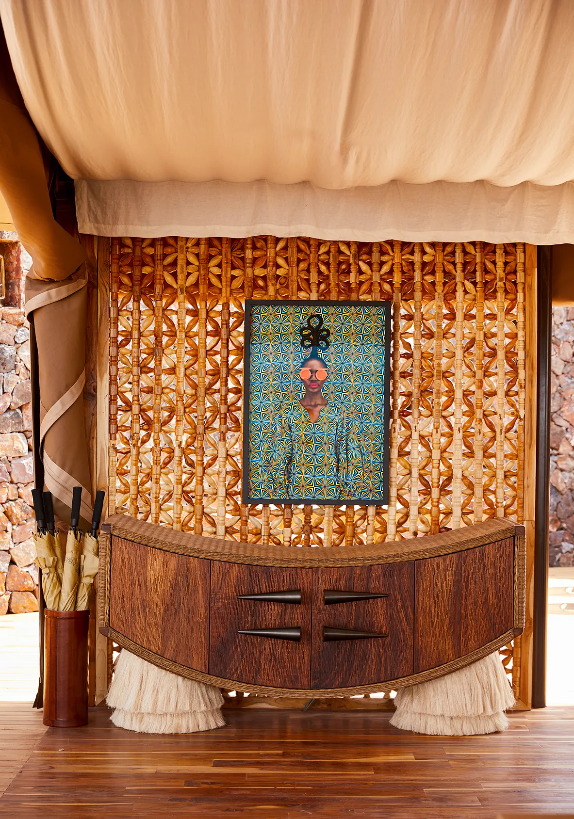 Curved wooden sideboard beneath a framed portrait, set against an intricate woven wall in a safari-style interior.