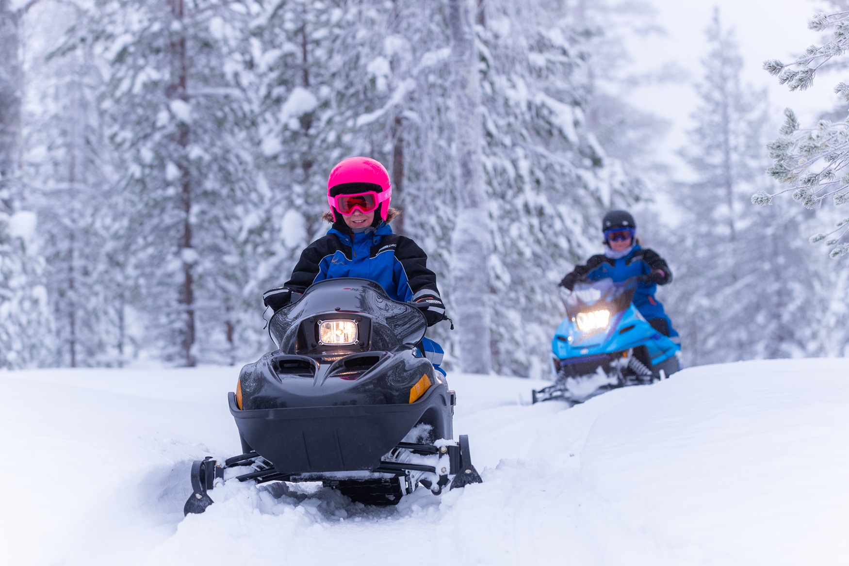 Two people wearing colourful snow gear riding snow mobiles through a wintery forest.