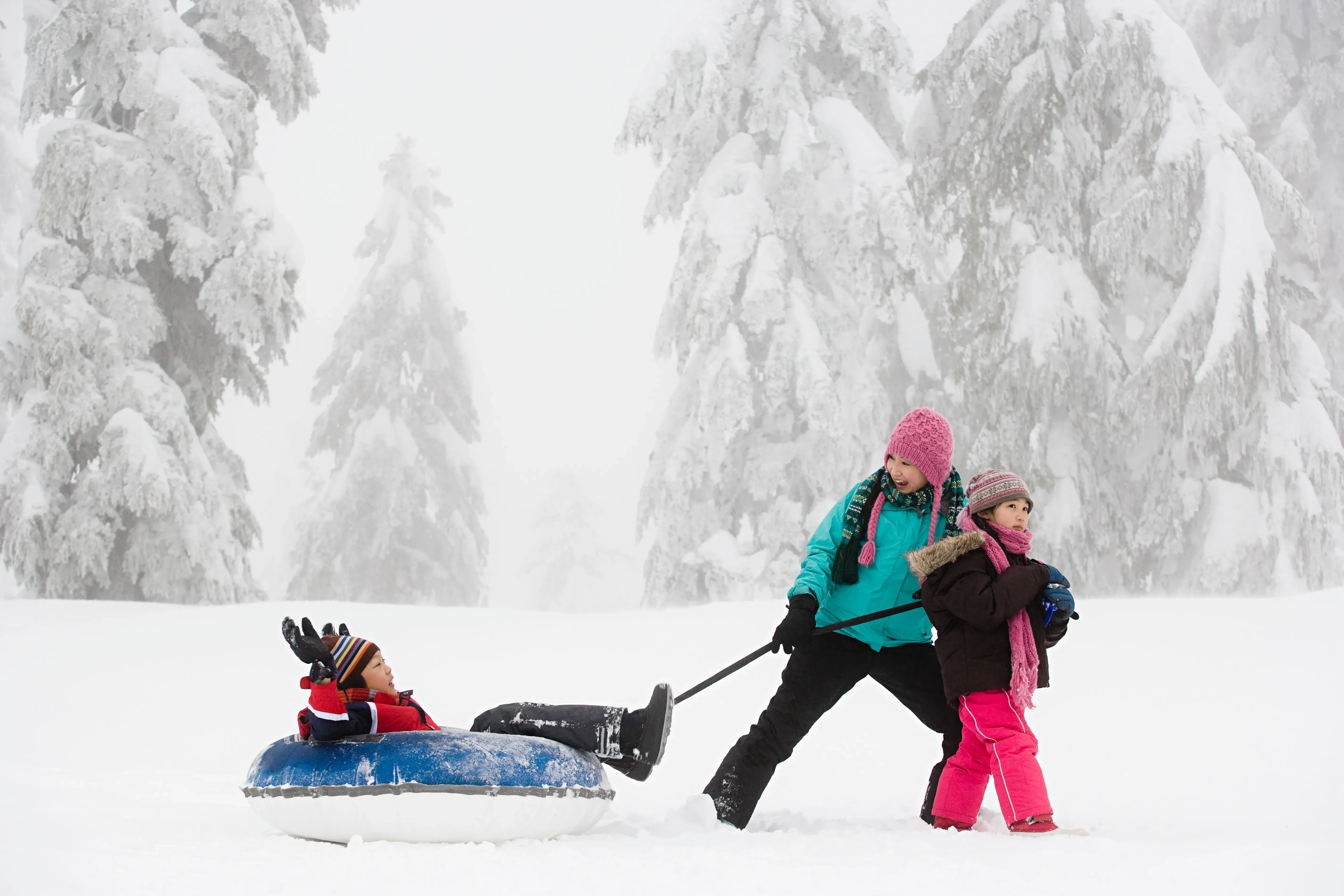 Three children playing in the snow with one child sat in a snow tube being pulled along by another