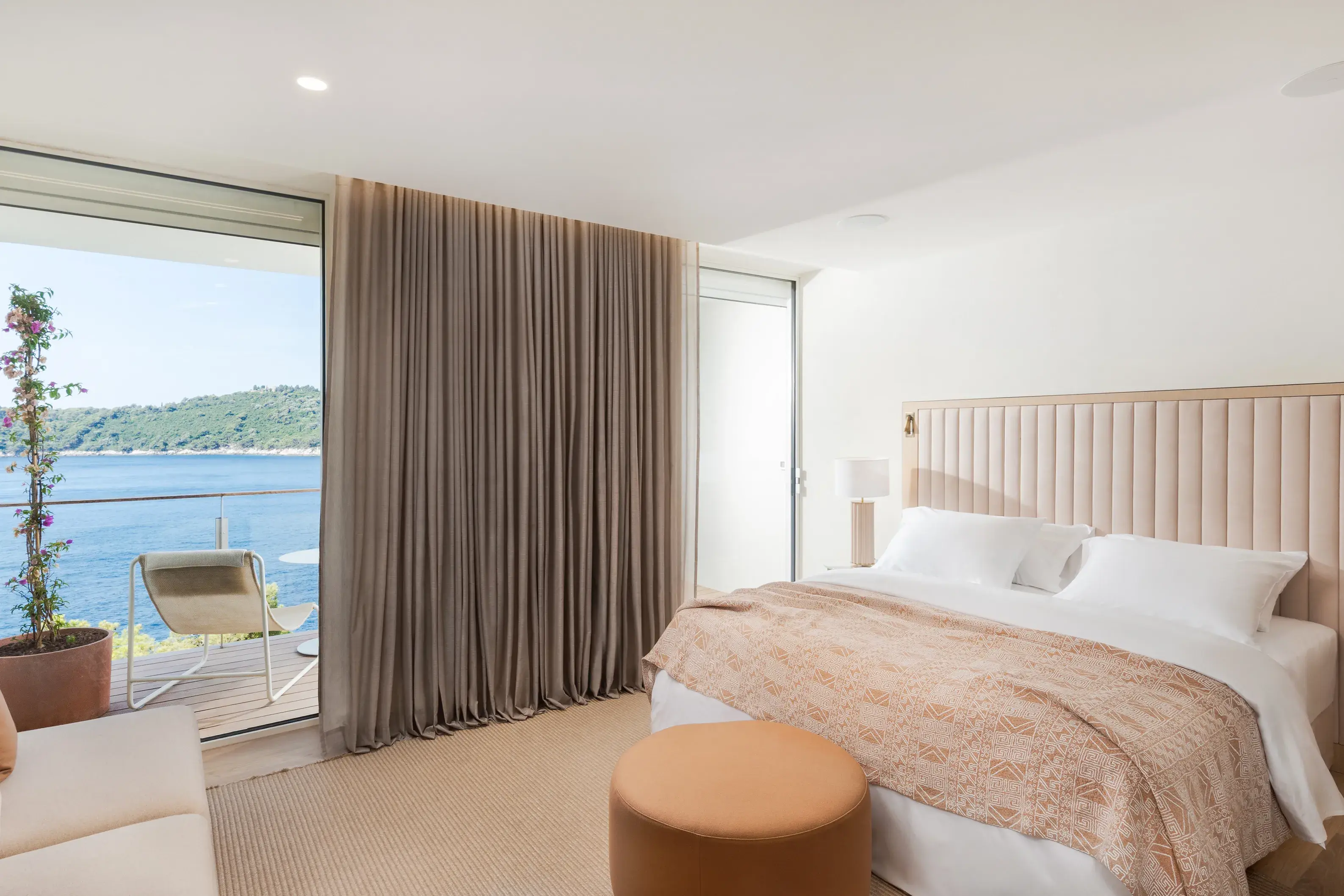Deluxe Sea View room at Villa Dubrovnik in Croatia featuring a comfy bed with pale orange soft furnishings and large balcony doors with curtains half drawn revealing a glimpse of the sea view and balcony furniture
