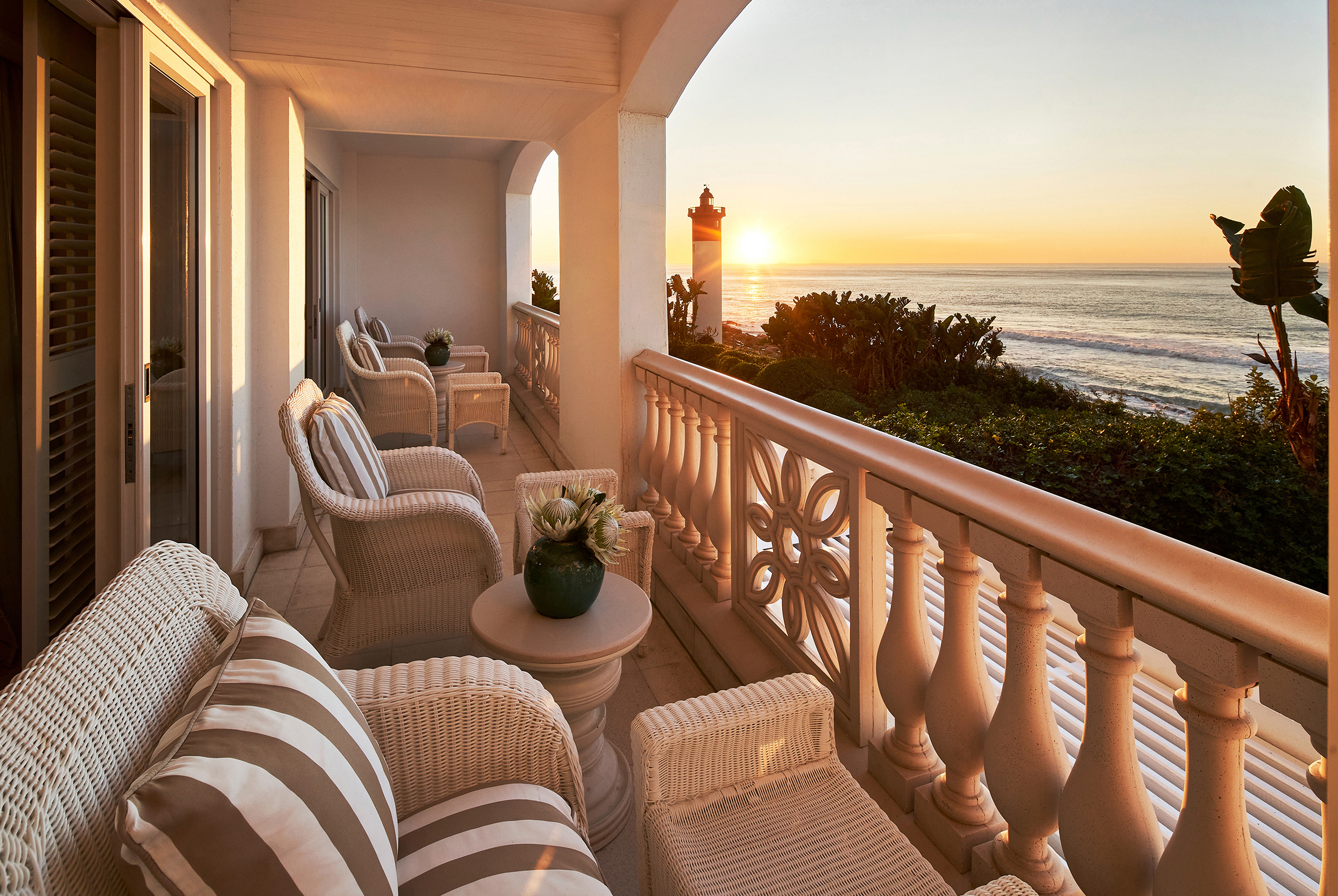 The terrace of a suite at The Oyster Box featuirng rattan and striped furniture with a sweeping view of the sea and lighthouse in the distance