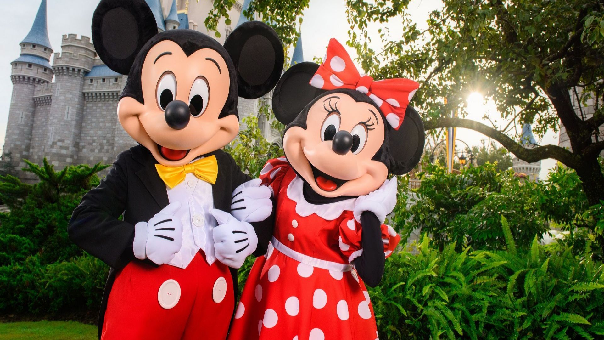  Mickey Mouse and Minnie Mouse posing together in front of a castle with greenery around them
