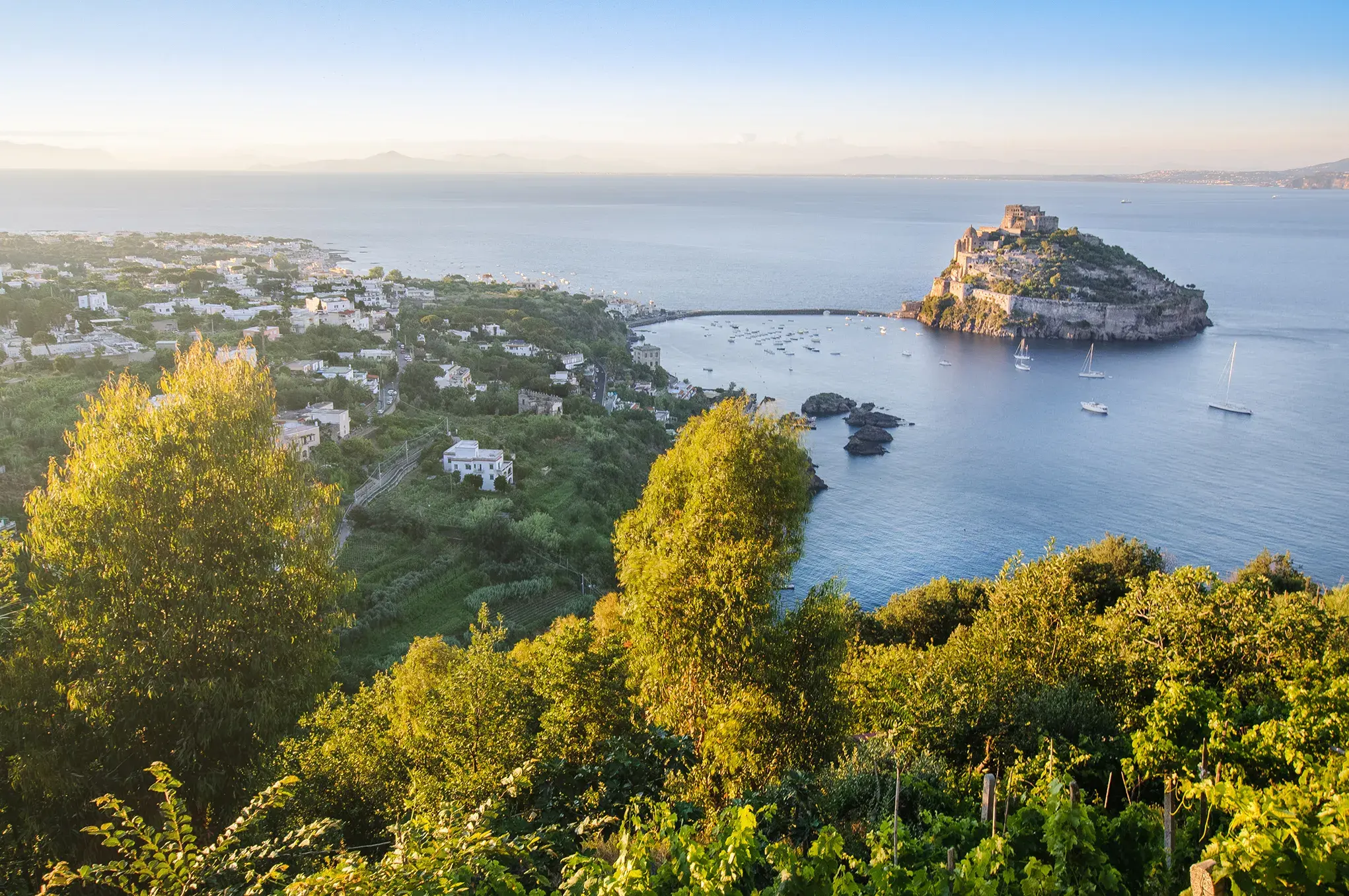 Panoramic view of Ischia island with Aragonese Castle surrounded by blue sea and lush green hills at sunset.