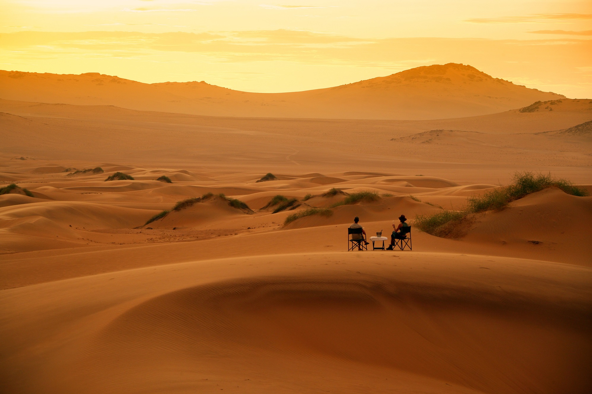 Two people sit on chairs in a vast desert landscape with sand dunes and hills in the background during sunset.