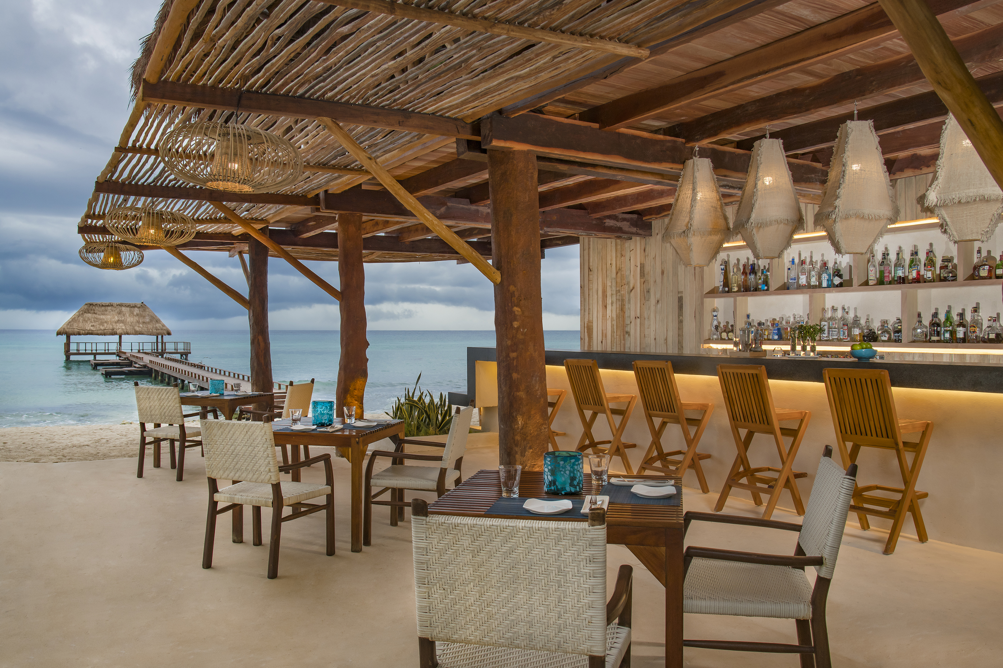 A thatched roof bar beside the sea with dining tables and stools and a pier in the background