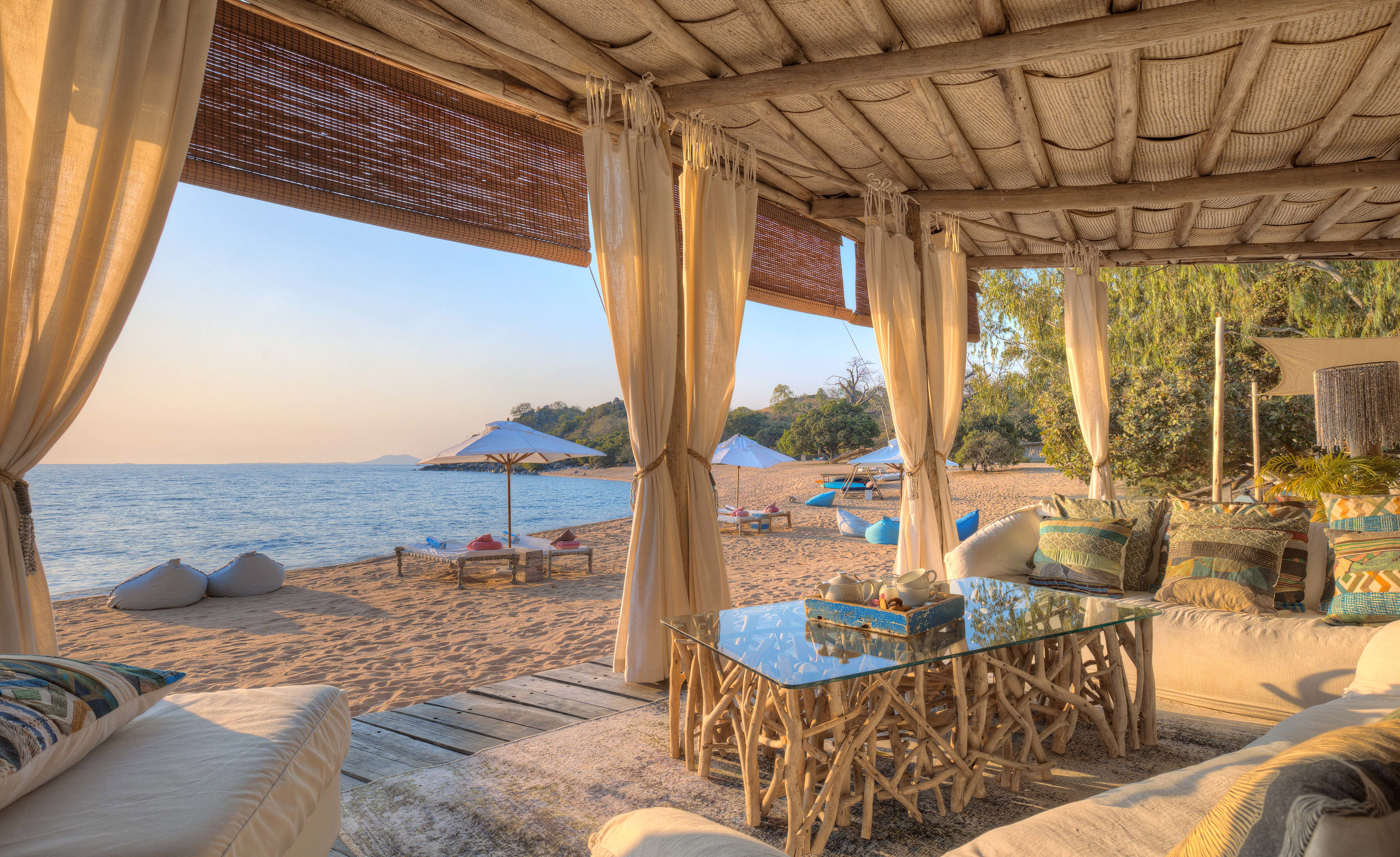 A luxurious beachside lounge area at Kaya Mawa with comfortable seating, a glass and driftwood table, and curtains framing a view of the sandy beach, sun loungers, umbrellas, and the calm blue waters of Likoma Island.