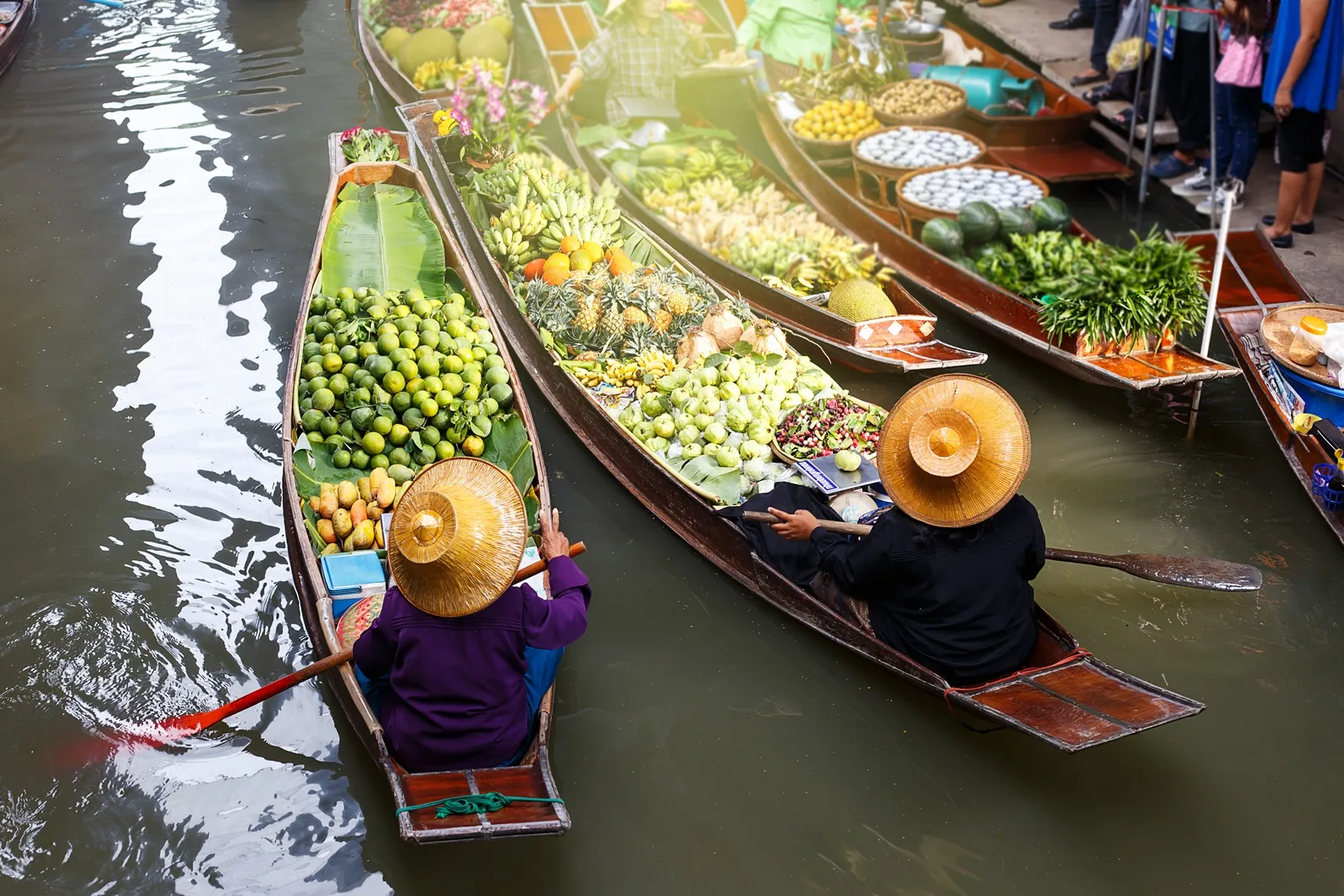 Two people in straw hats paddle wooden boats filled with fruits and vegetables on a canal.