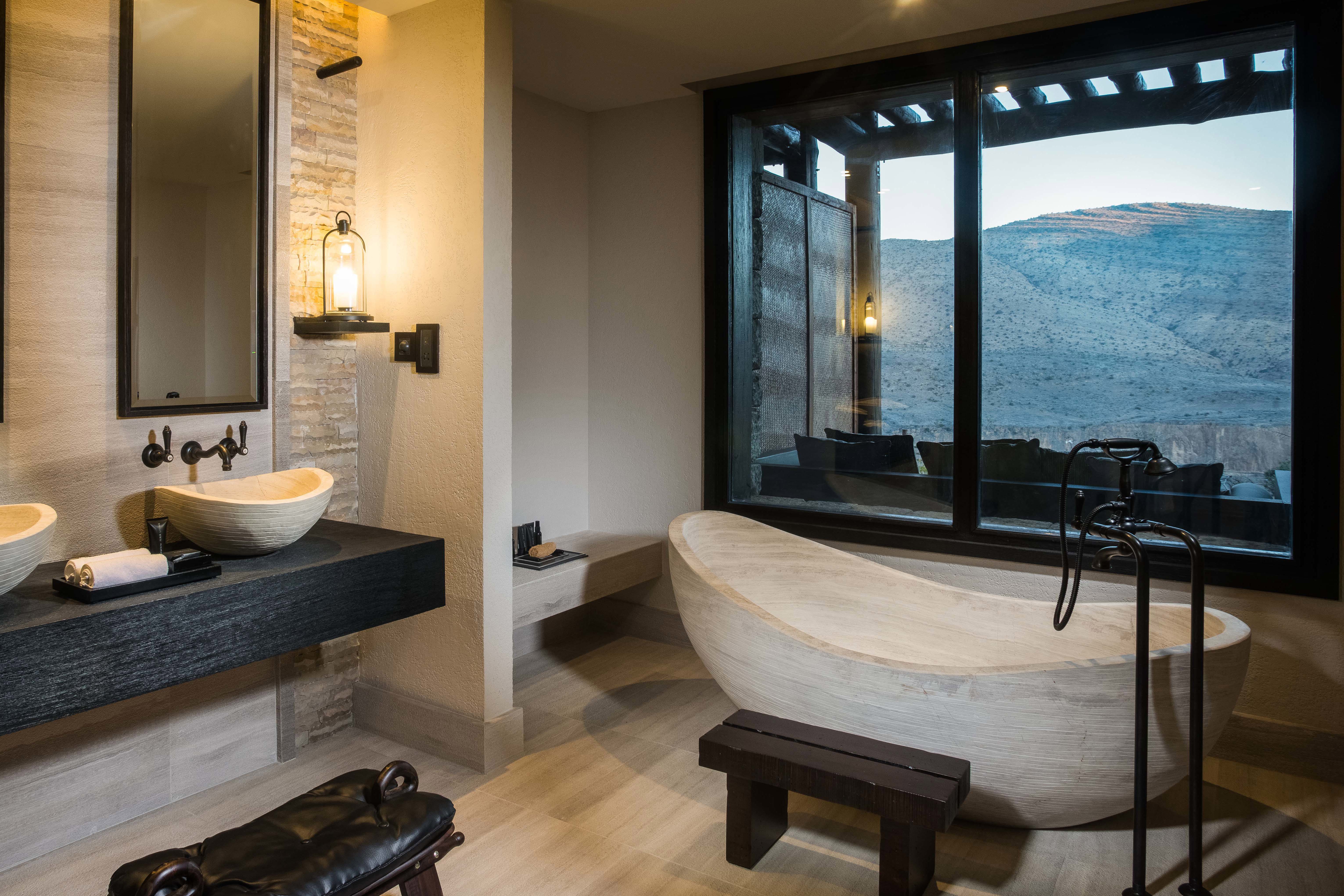 A modern stone bathroom at Alila Jabal Akhdar featuring a large stand-alone tub and black fixtures, with a large window looking out over mountains