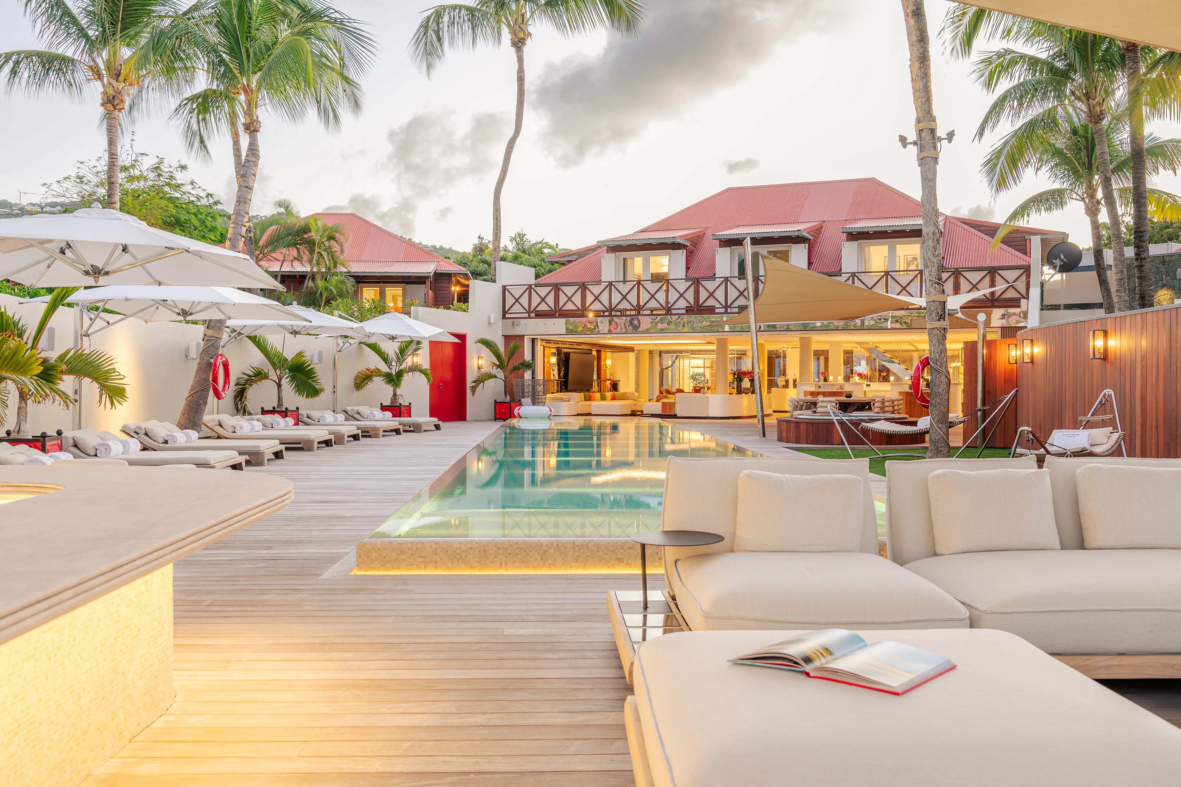 The outdoor terrace of Villa Rockstar at Eden Rock in St Barths featuring comfy sun beds, white sun umbrellas and a pool set before a red-roofed villa