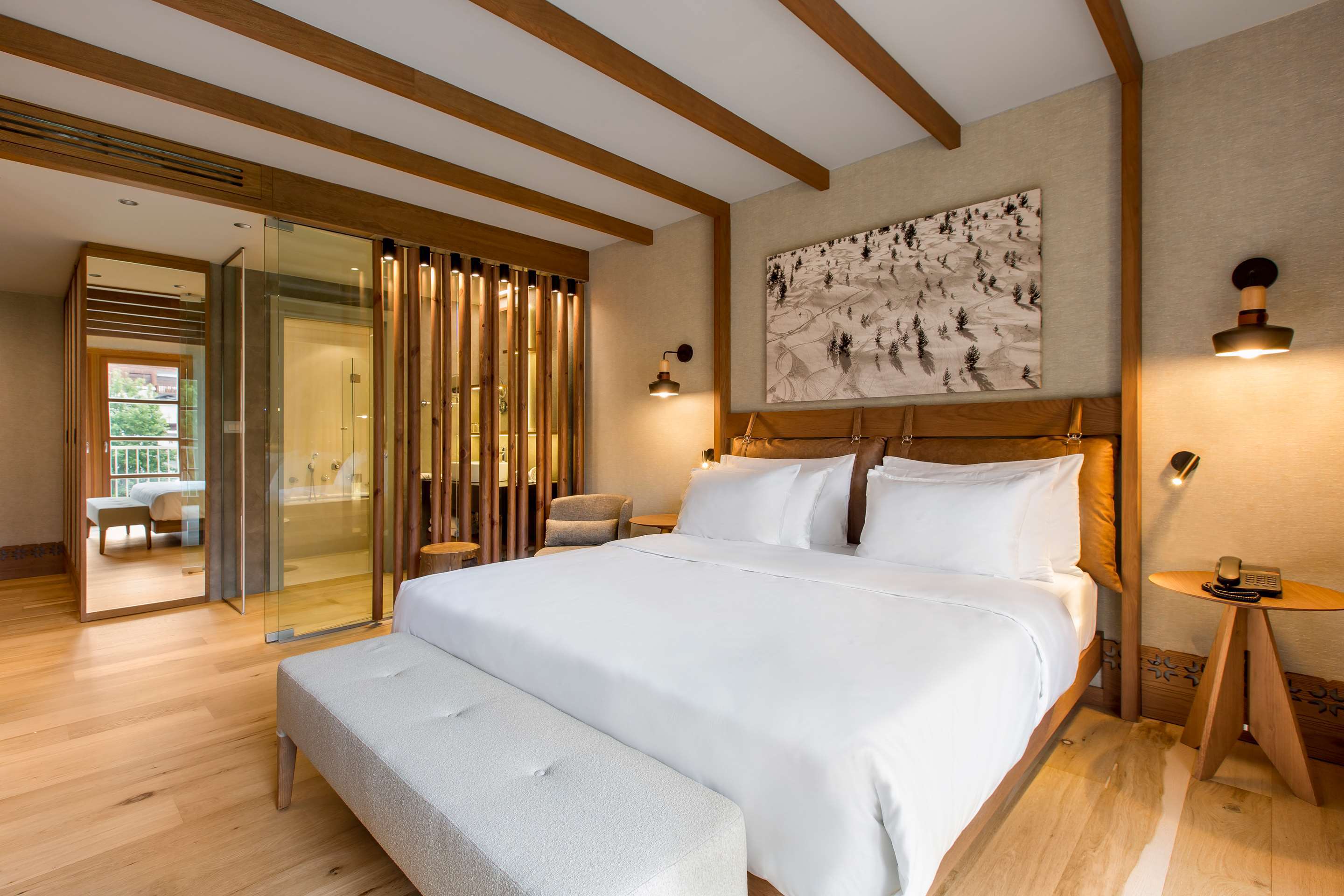 A cosy beamed bedroom featuring a bed with Alpine artwork, ottoman and a glimpse of the bathroom beyond