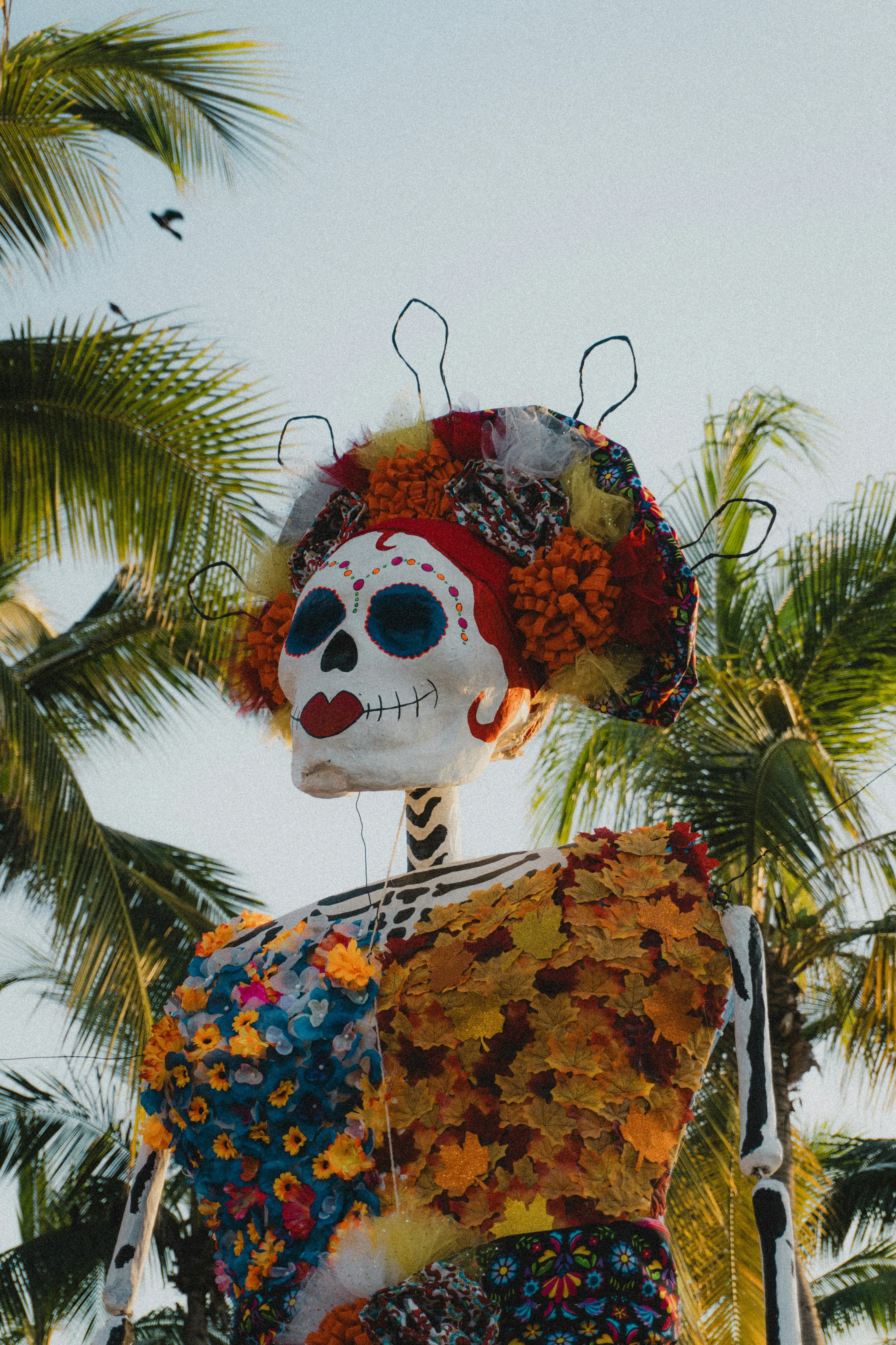 Colourful skeleton figure for Mexico's Day of the Dead decorated with vibrant flowers and wire details, set against palm trees and a clear sky.