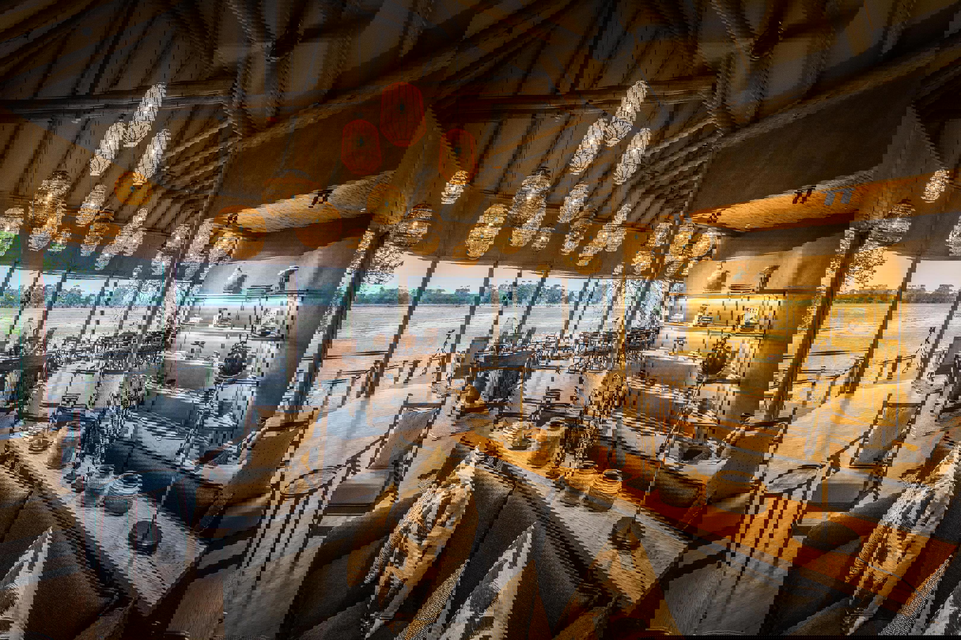 Interior of Puku Ridge Camp showcasing a spacious lounge with modern decor, wooden furniture, and large windows overlooking the landscape, illuminated by hanging lanterns for a warm ambiance