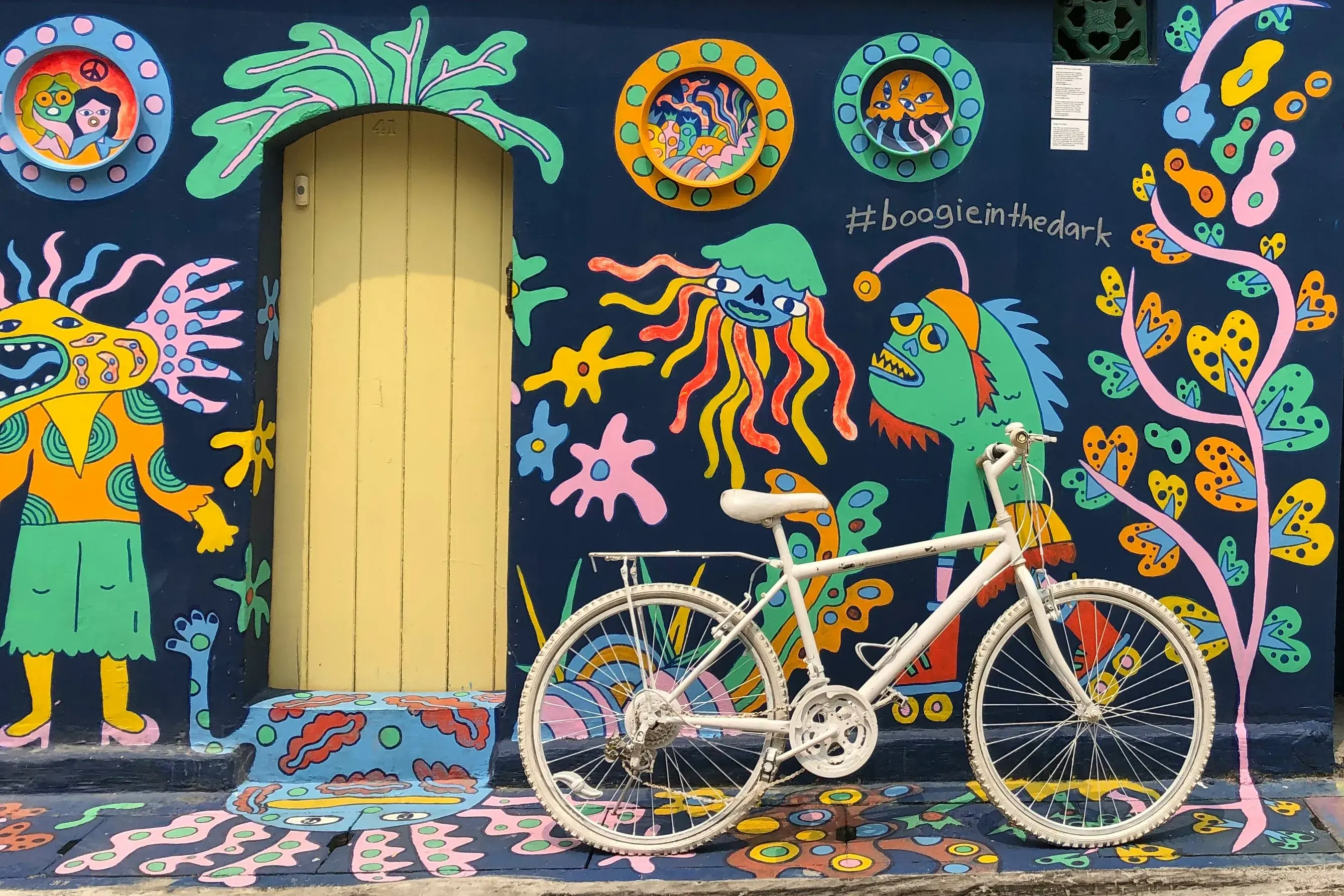 A white bicycle parked outside blue concrete wall filled with multicoloured street art