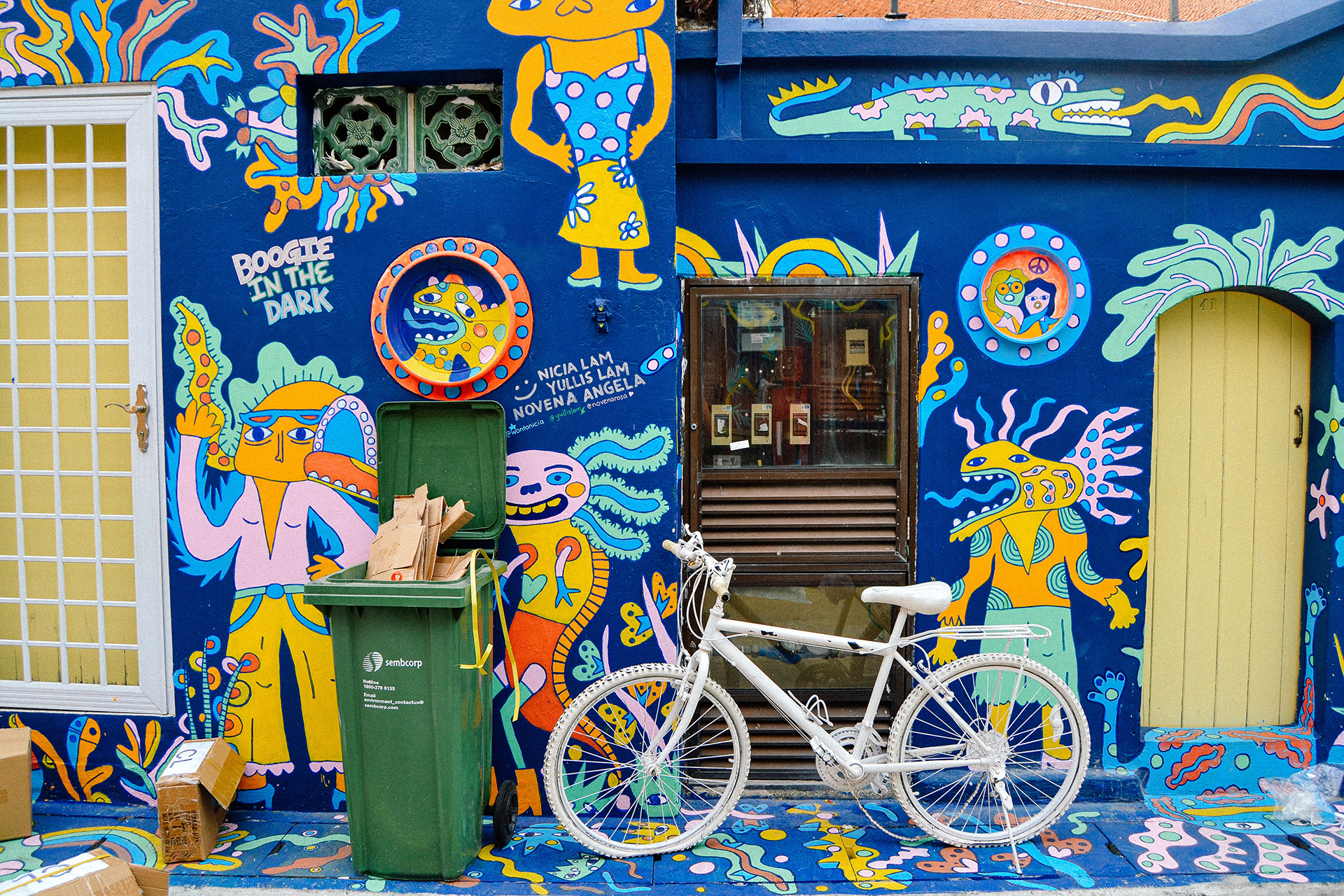 A white bicycle parked outside blue concrete wall filled with multicoloured street art