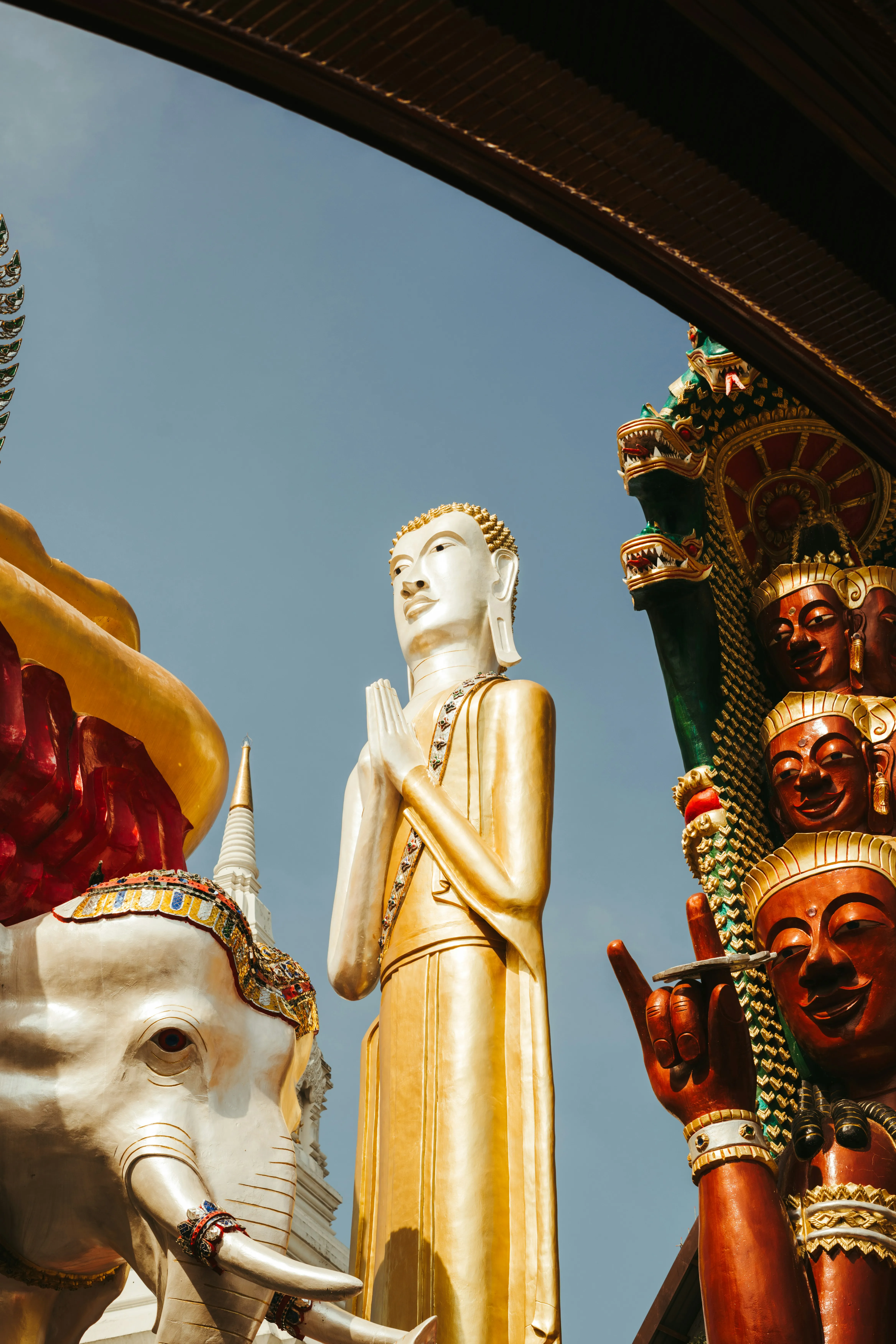 Golden Buddha statue with hands in prayer position surrounded by ornate temple decorations and an elephant sculpture.