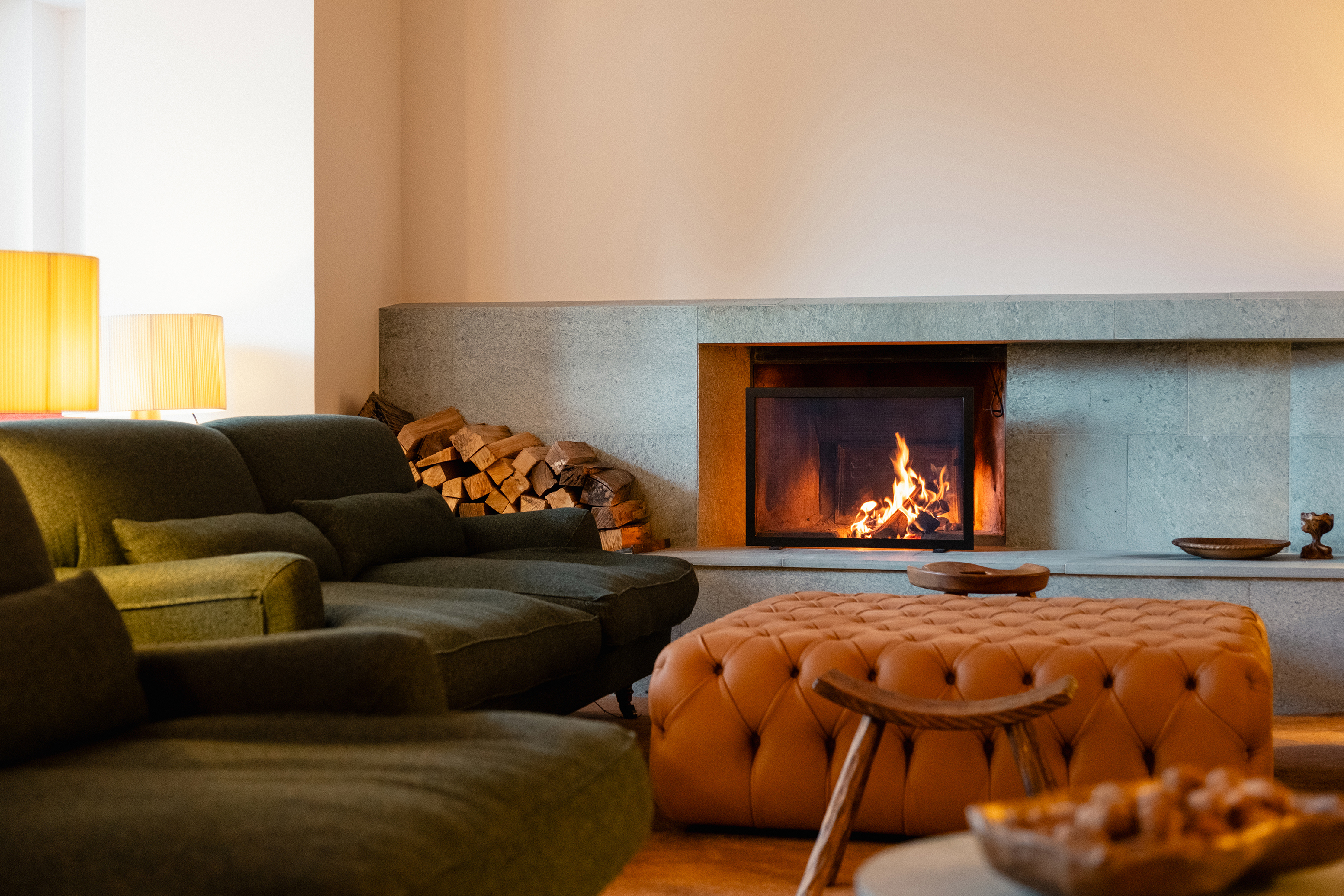 A cosy lobby featuring comfy green and orange sofa furniture in front of a lit fireplace with logs beside it